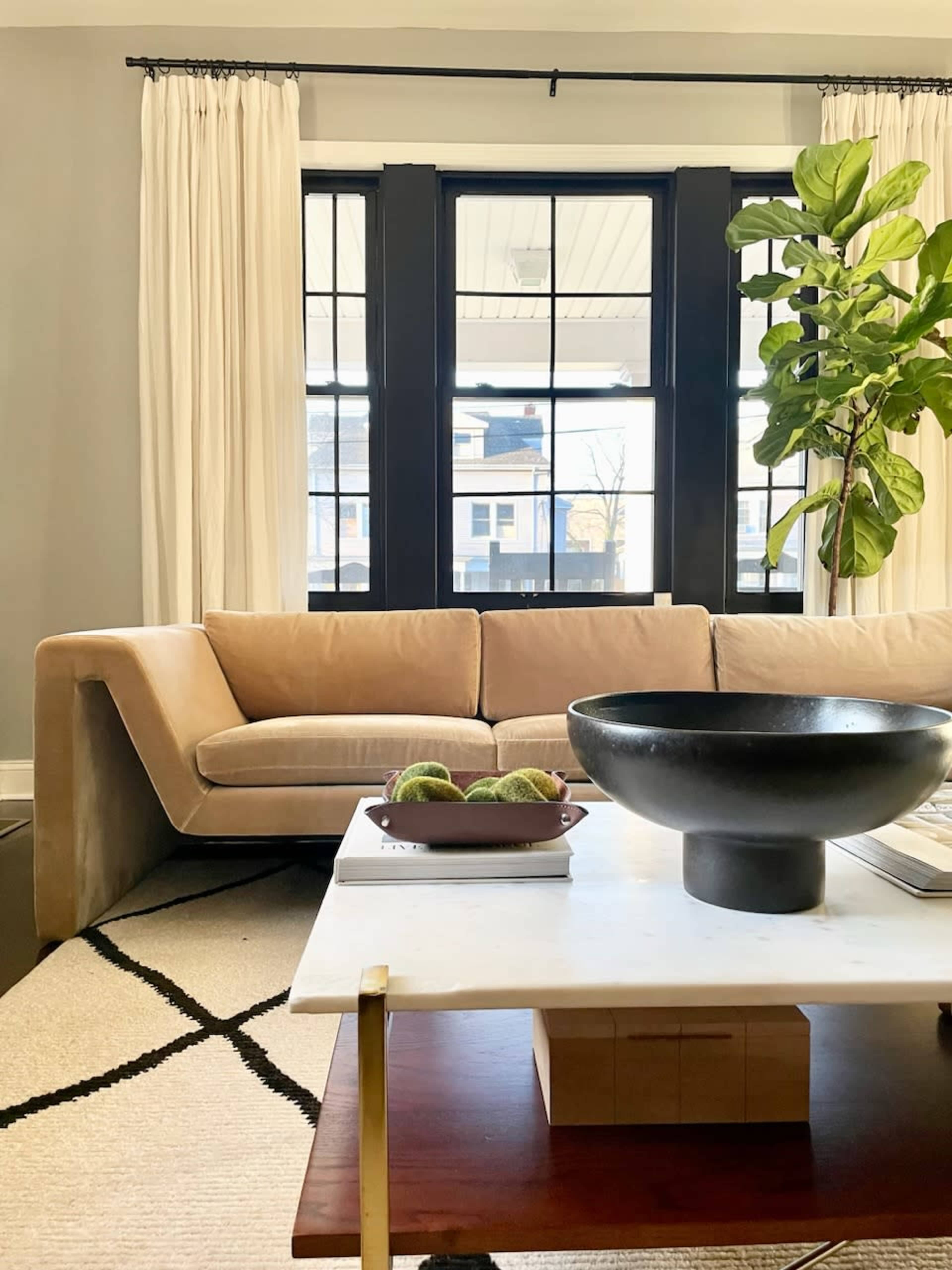 A modern living room features a curved tan sofa, a wooden coffee table with a marble top, and a large black bowl with green fruit, alongside a tall potted plant and large windows.