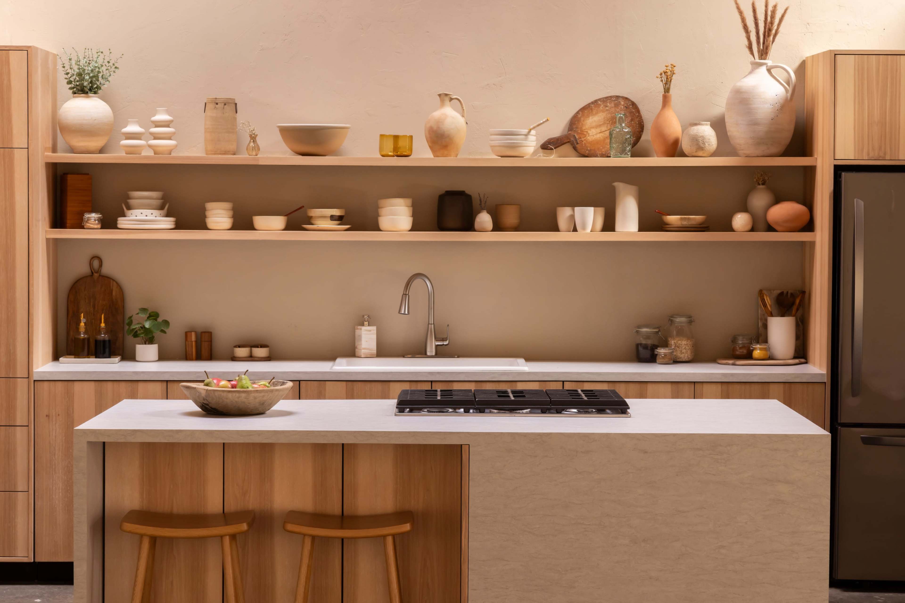 The image shows a modern kitchen with light wood cabinets, open shelving displaying various kitchenware, and a central island with two wooden stools.