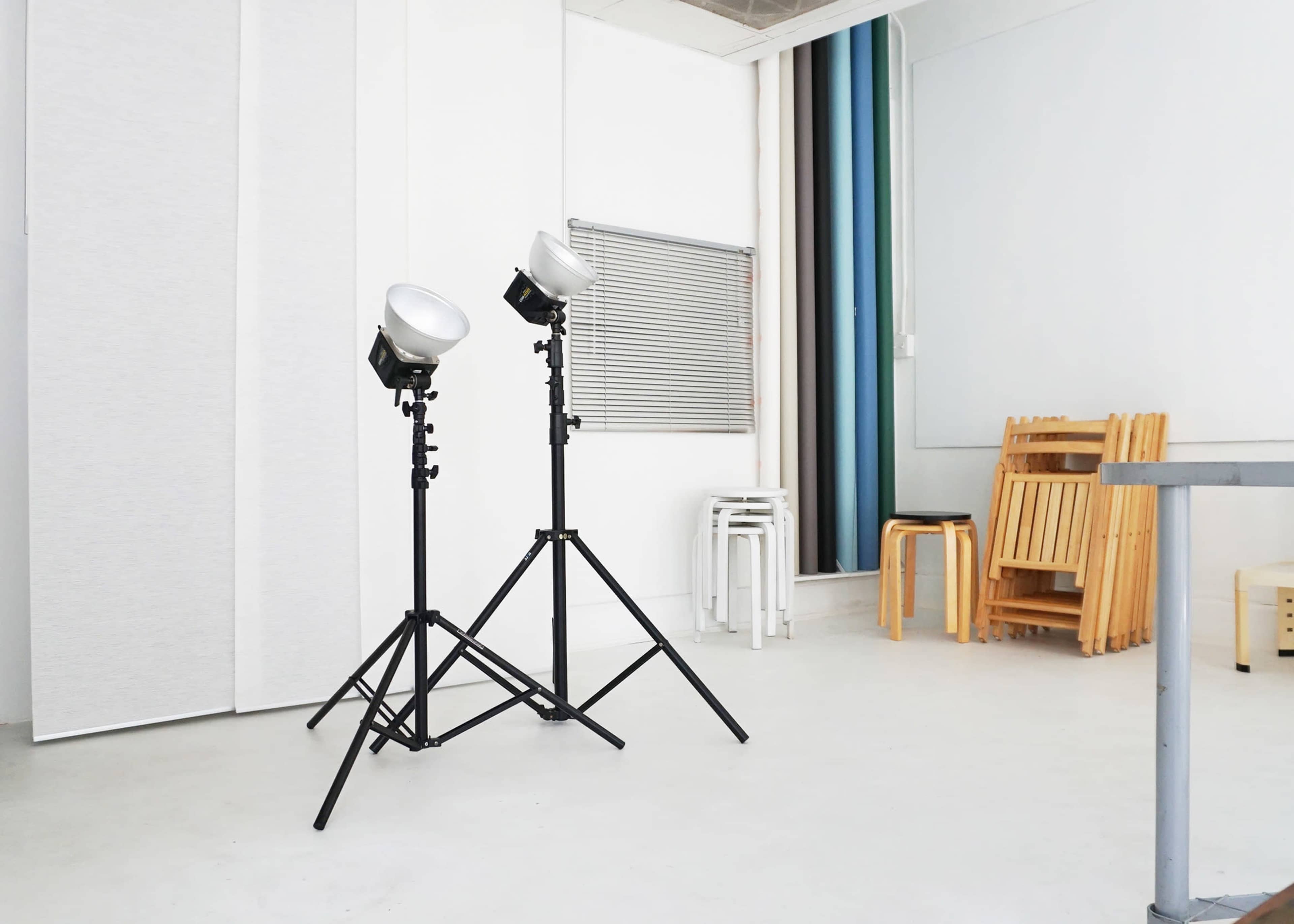 Two studio lights on tripods positioned in a minimalist room with a few chairs stacked against a wall.