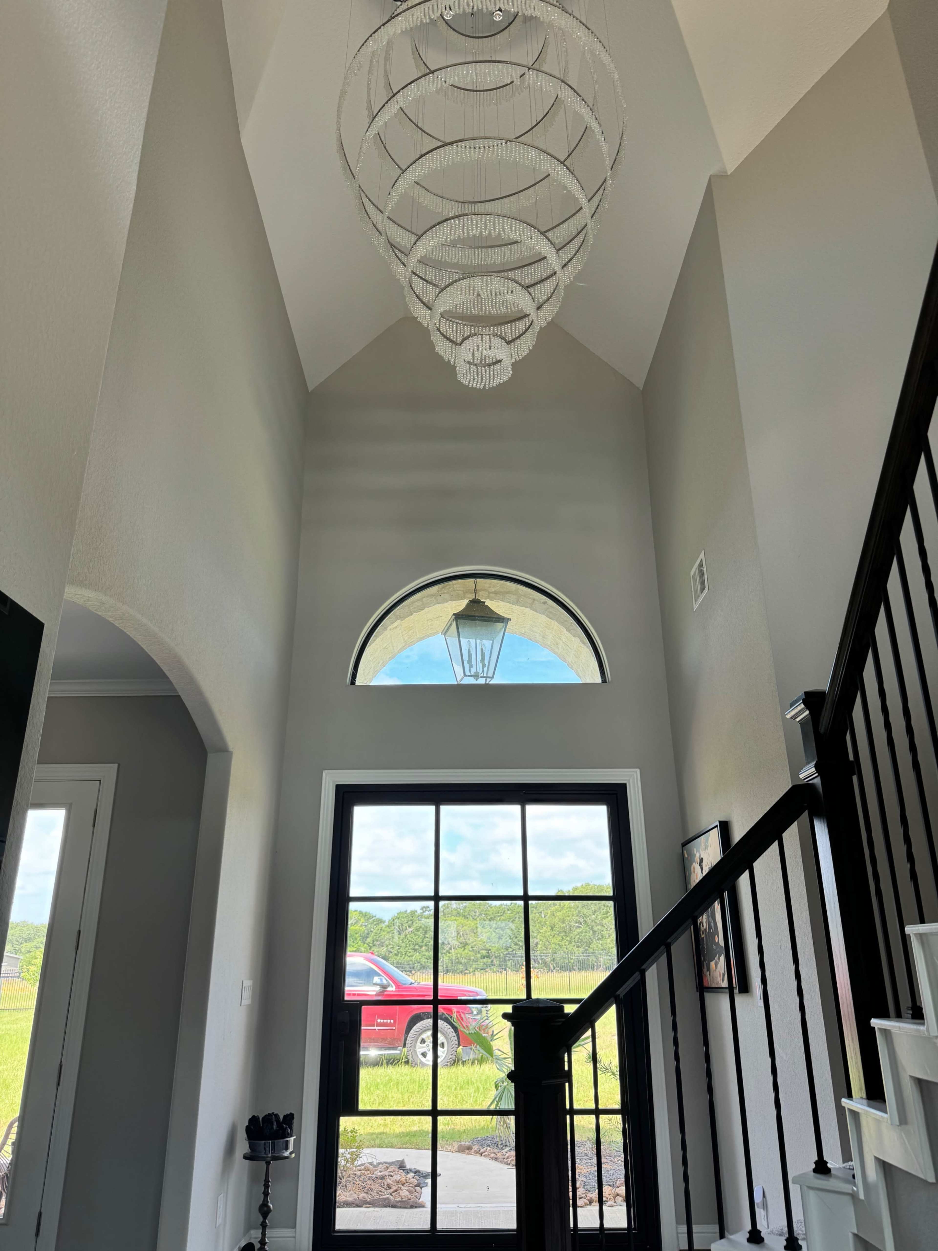 The image shows a two-story foyer with a large chandelier hanging from a vaulted ceiling and a view through a black-framed window of a red vehicle parked outside.