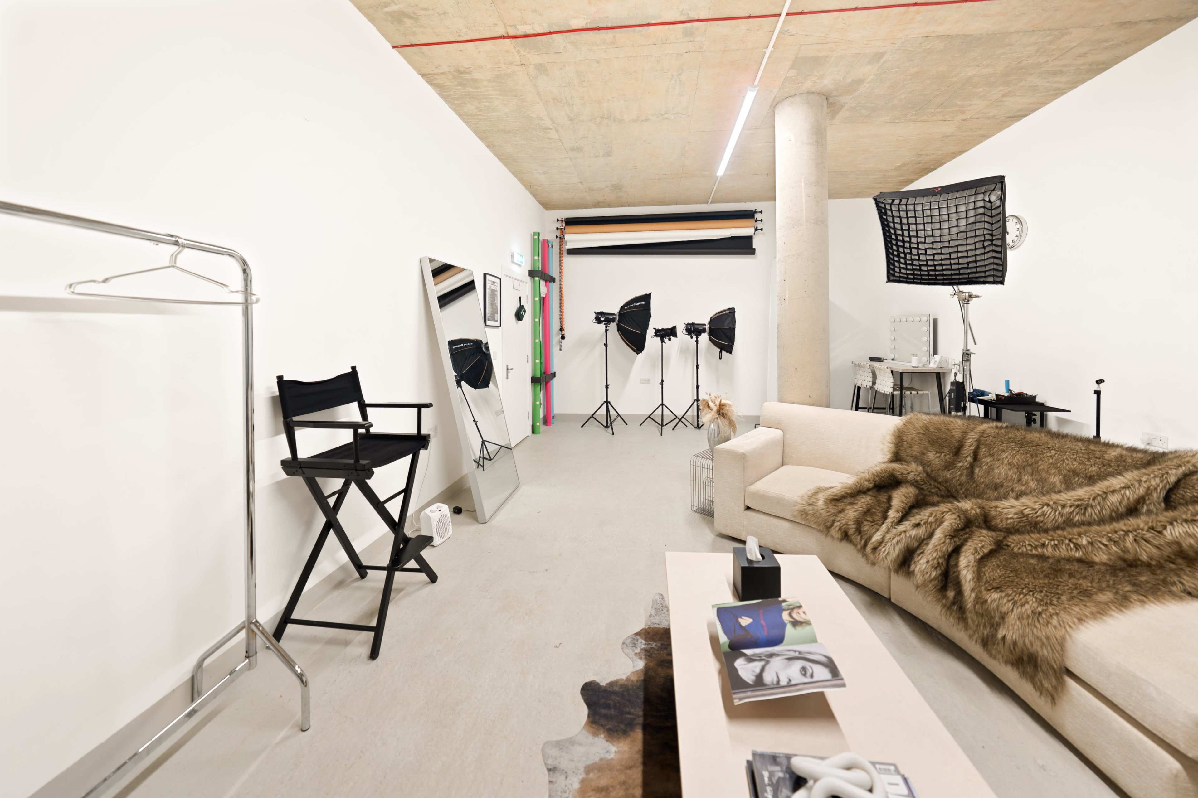 The image shows a spacious studio with a modern couch, a clothing rack, a large mirror, and multiple lighting equipment set up for photography.