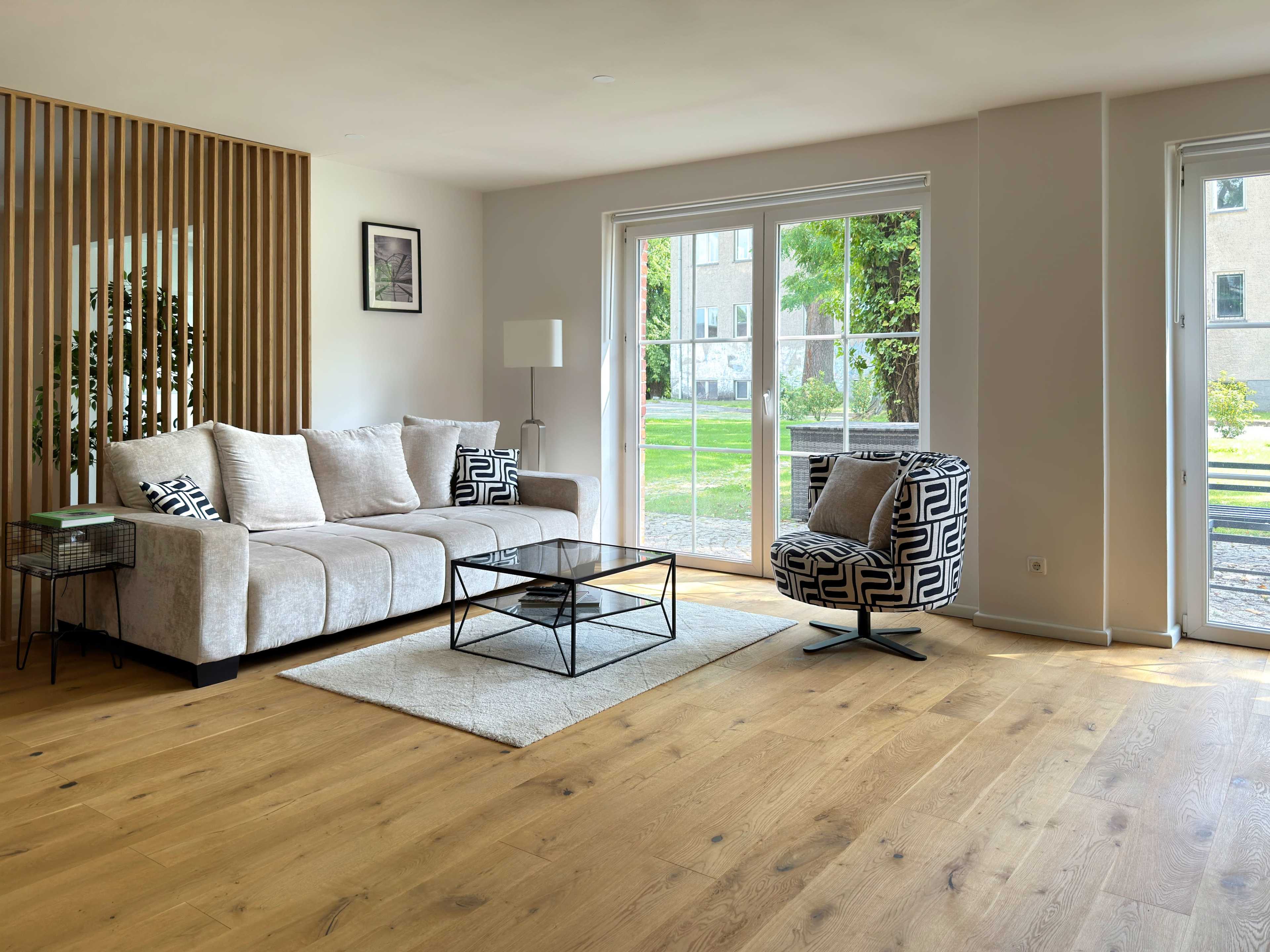 The living room features a light-colored sofa, a patterned armchair, a glass coffee table, and large windows that overlook a garden.