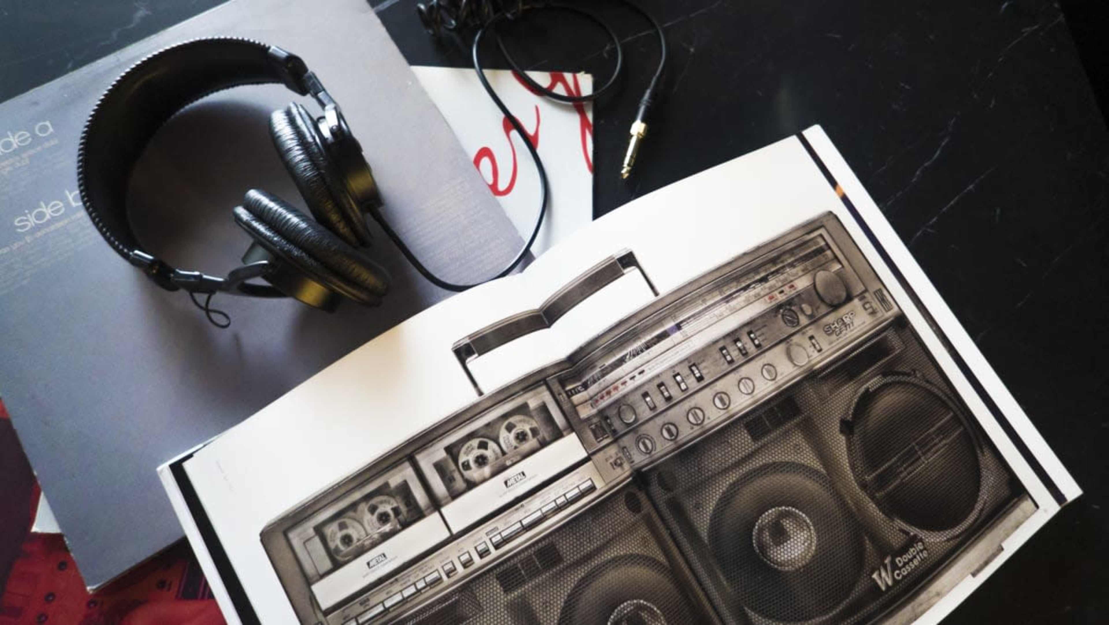 A pair of headphones rests on an open magazine featuring a large image of a vintage boombox, alongside a vinyl record and audio cable.