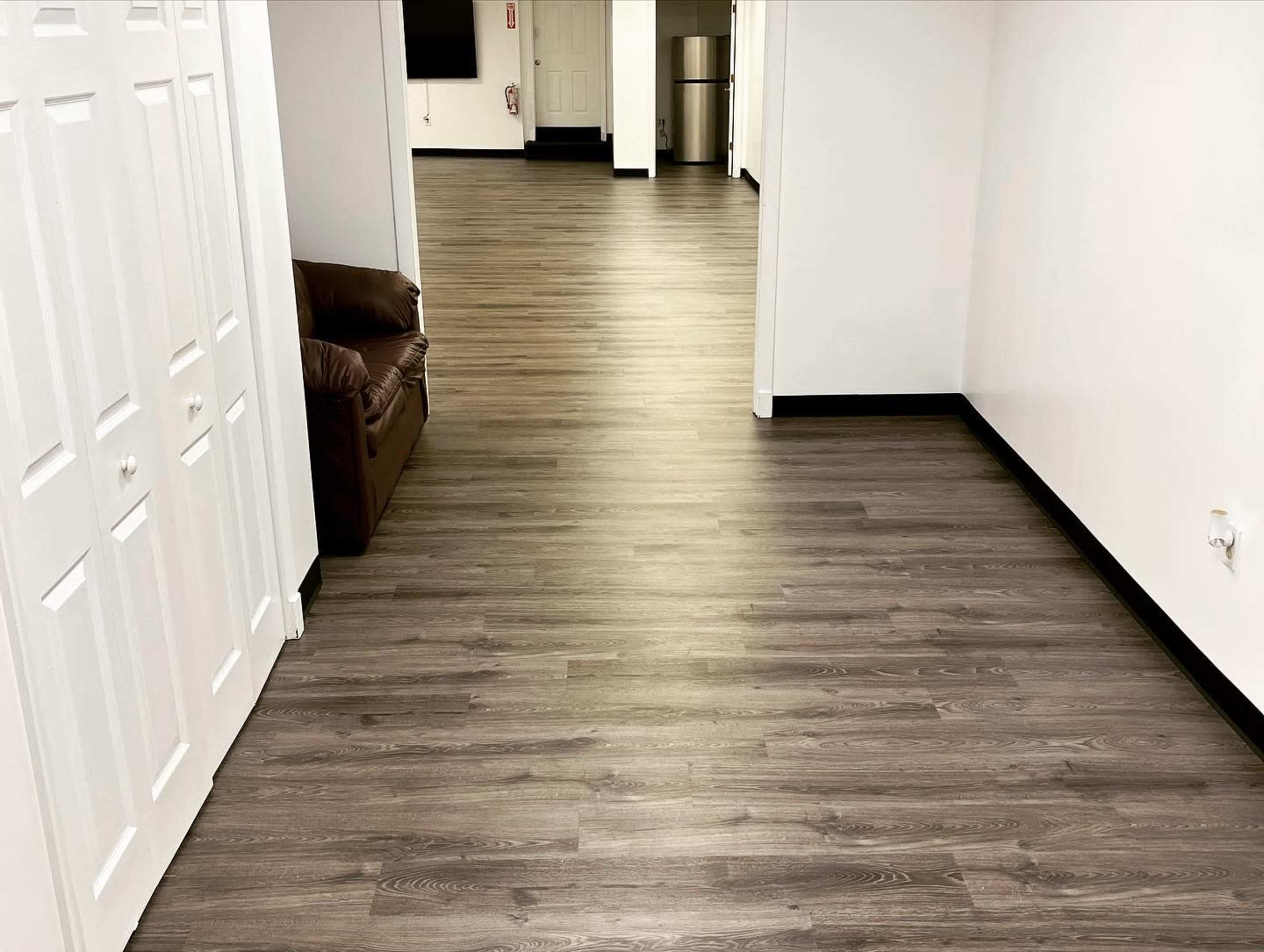 A hallway with laminate flooring, white walls, and a brown couch on one side.