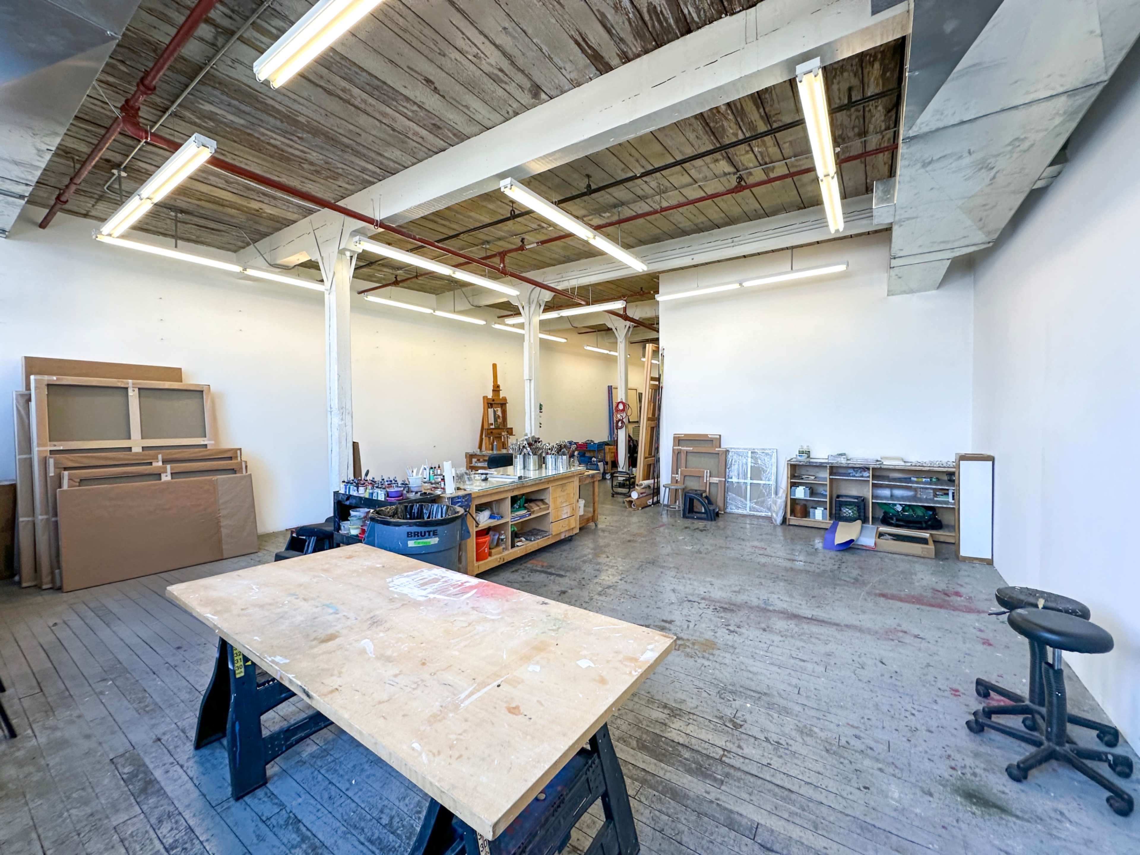 The image shows a spacious, well-lit art studio with wooden floors, a central table, and various tools and materials organized on shelves.