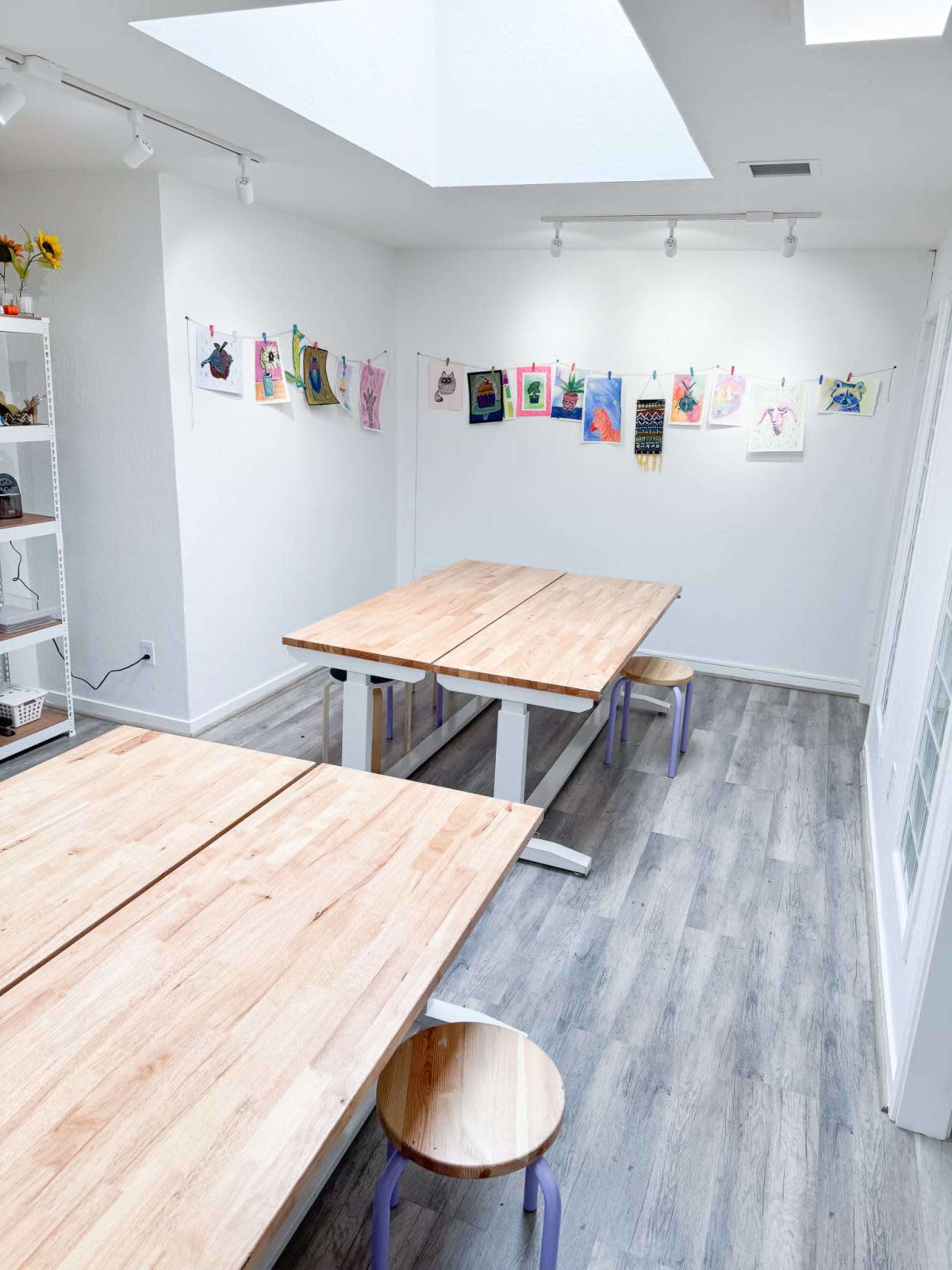 A well-lit art studio featuring two wooden tables and colorful artwork displayed on a wall.