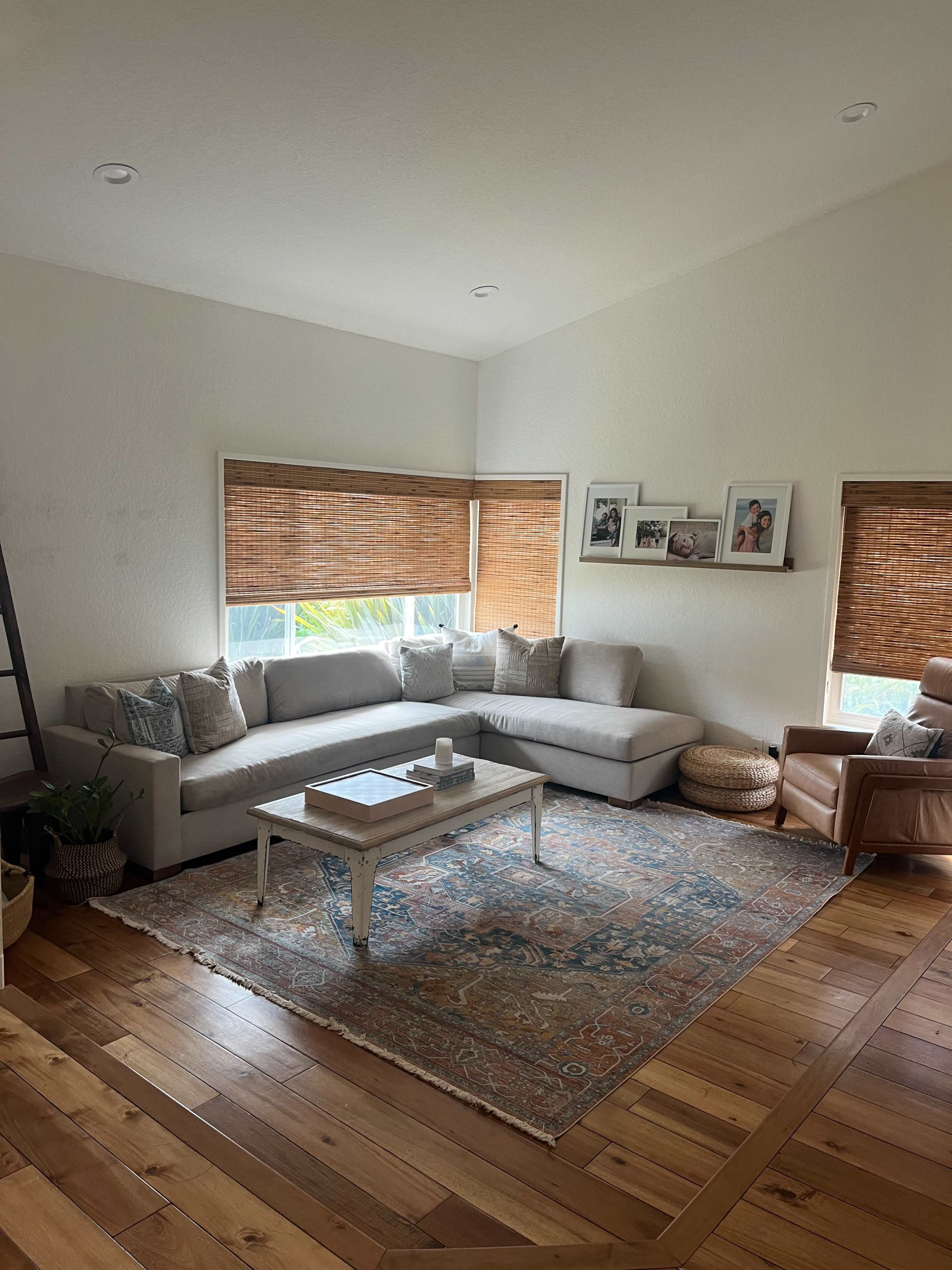 A light-filled living room features a grey sectional sofa, a wooden coffee table, and a large area rug, with bamboo shades covering the windows.