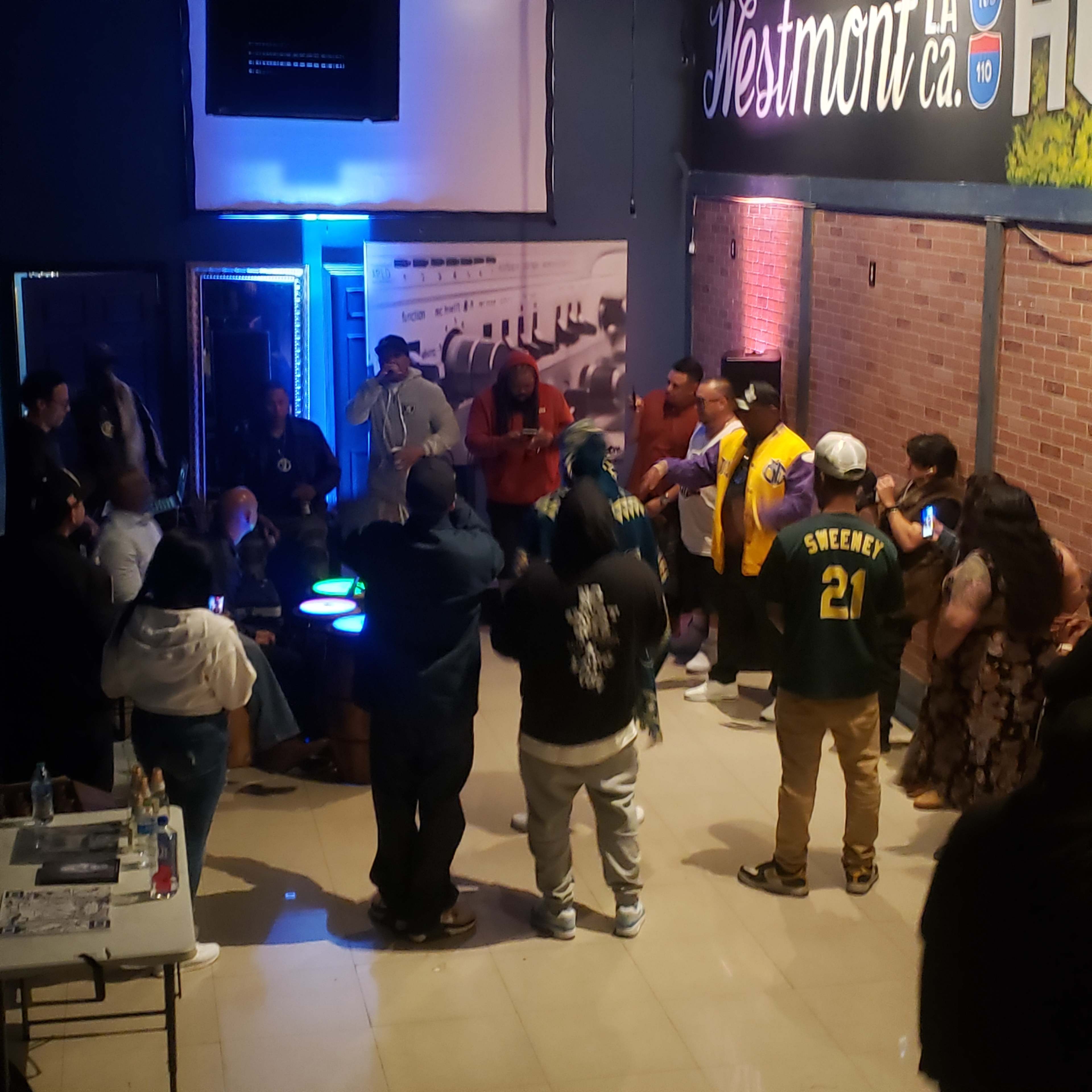 A group of people gathers in a dimly lit venue with brick walls, engaging in conversation and activities around circular tables.