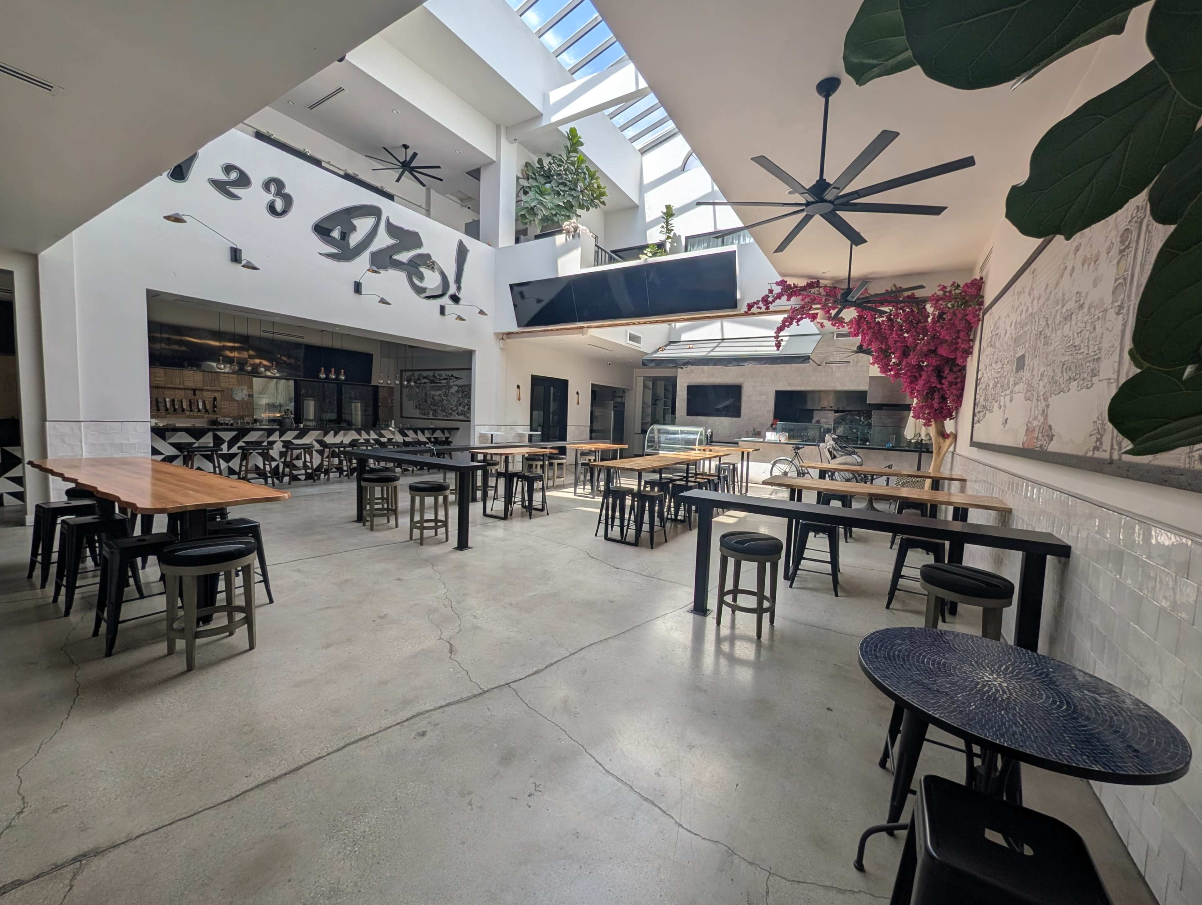 The image shows a spacious, well-lit restaurant interior with wooden tables and black stools arranged in an open layout, featuring large windows and greenery.