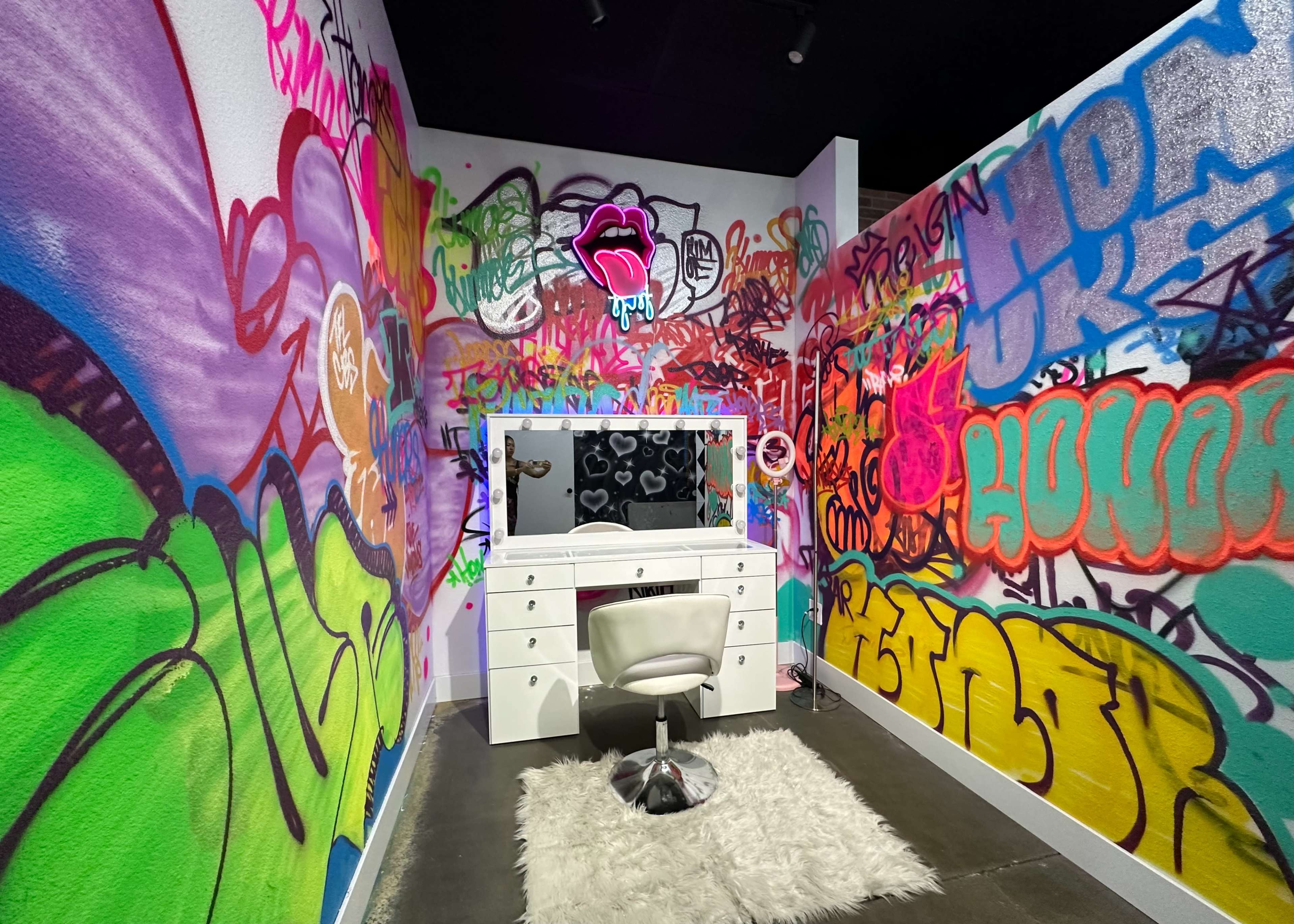A brightly colored room with graffiti-covered walls, featuring a white vanity with a mirror and a chair on a fluffy rug.