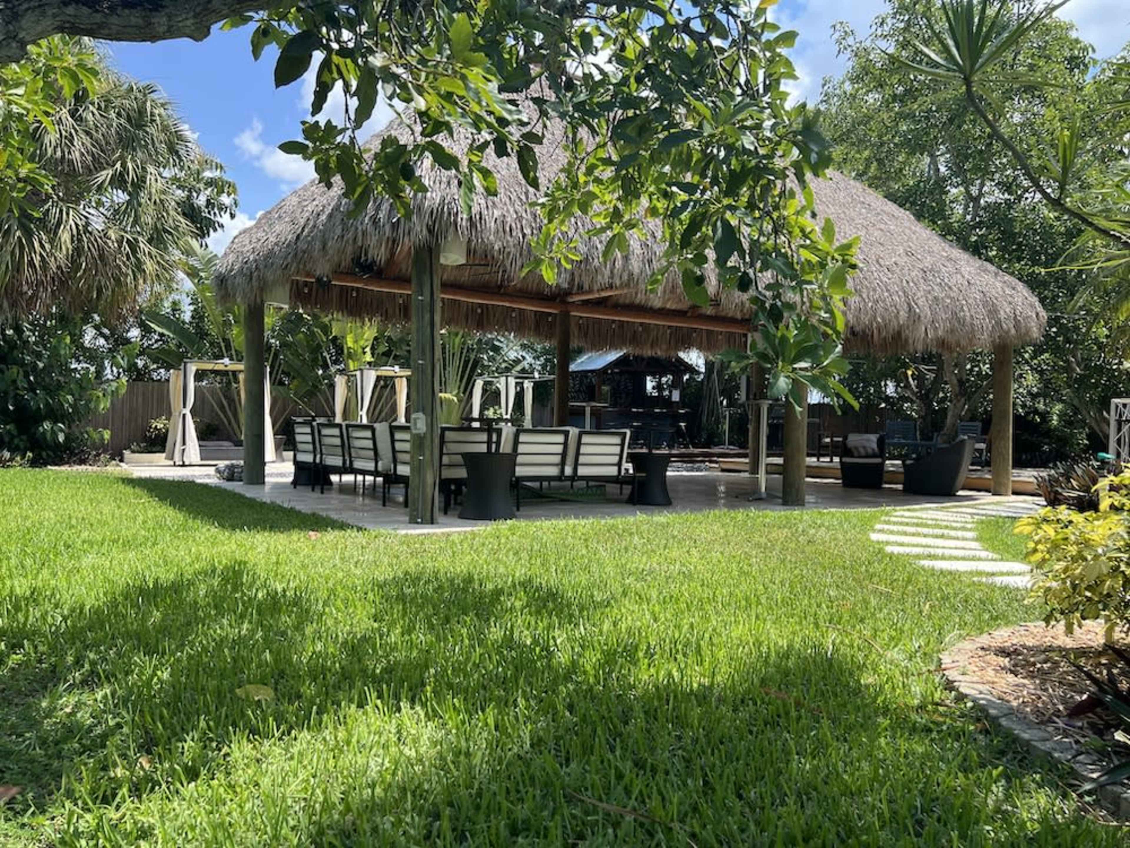 A shaded outdoor pavilion with a thatched roof contains a dining area surrounded by lush greenery and a well-maintained lawn.