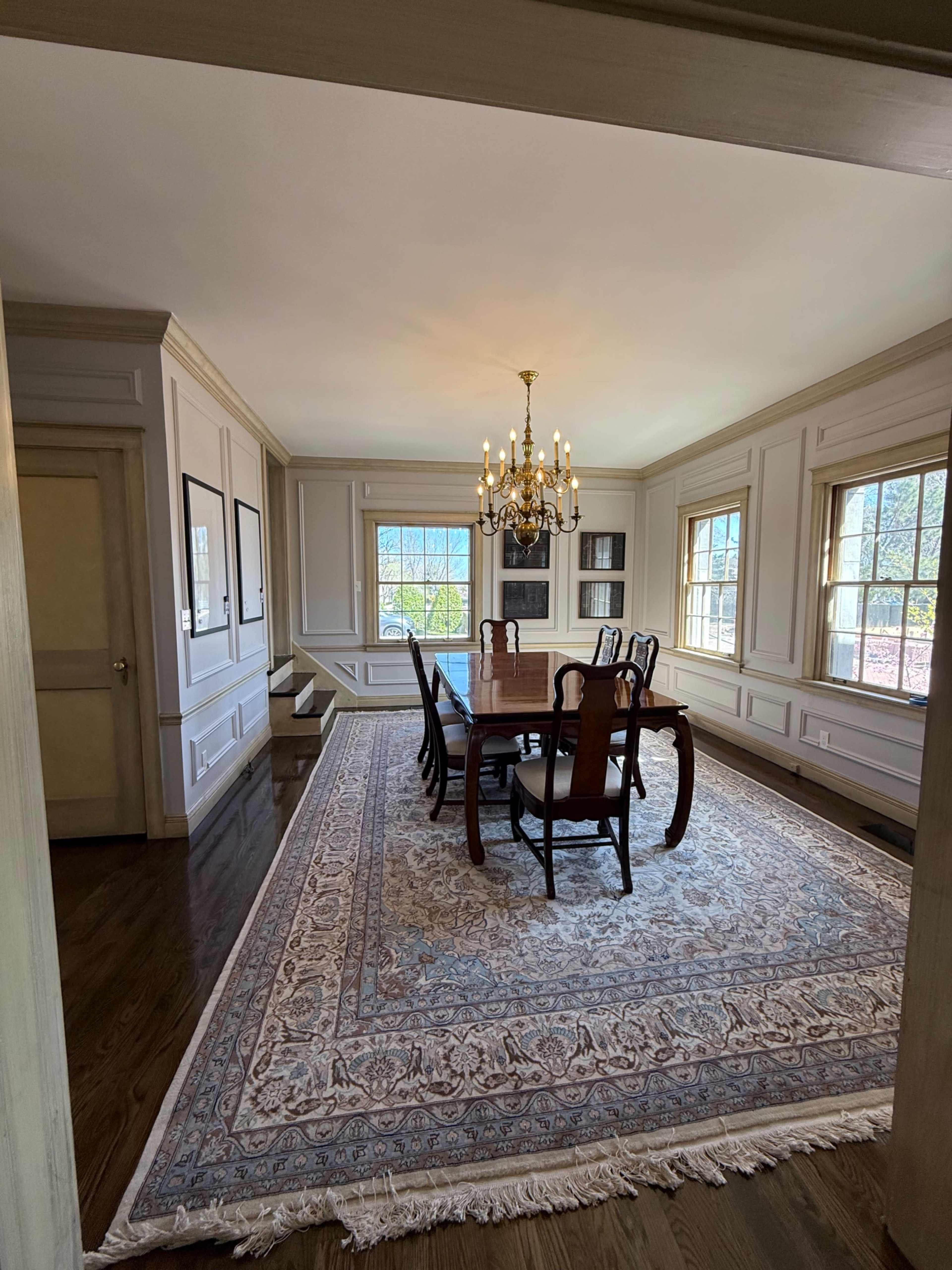 The image shows a spacious dining room featuring a large wooden table surrounded by chairs, a chandelier overhead, and large windows allowing natural light to enter the space.