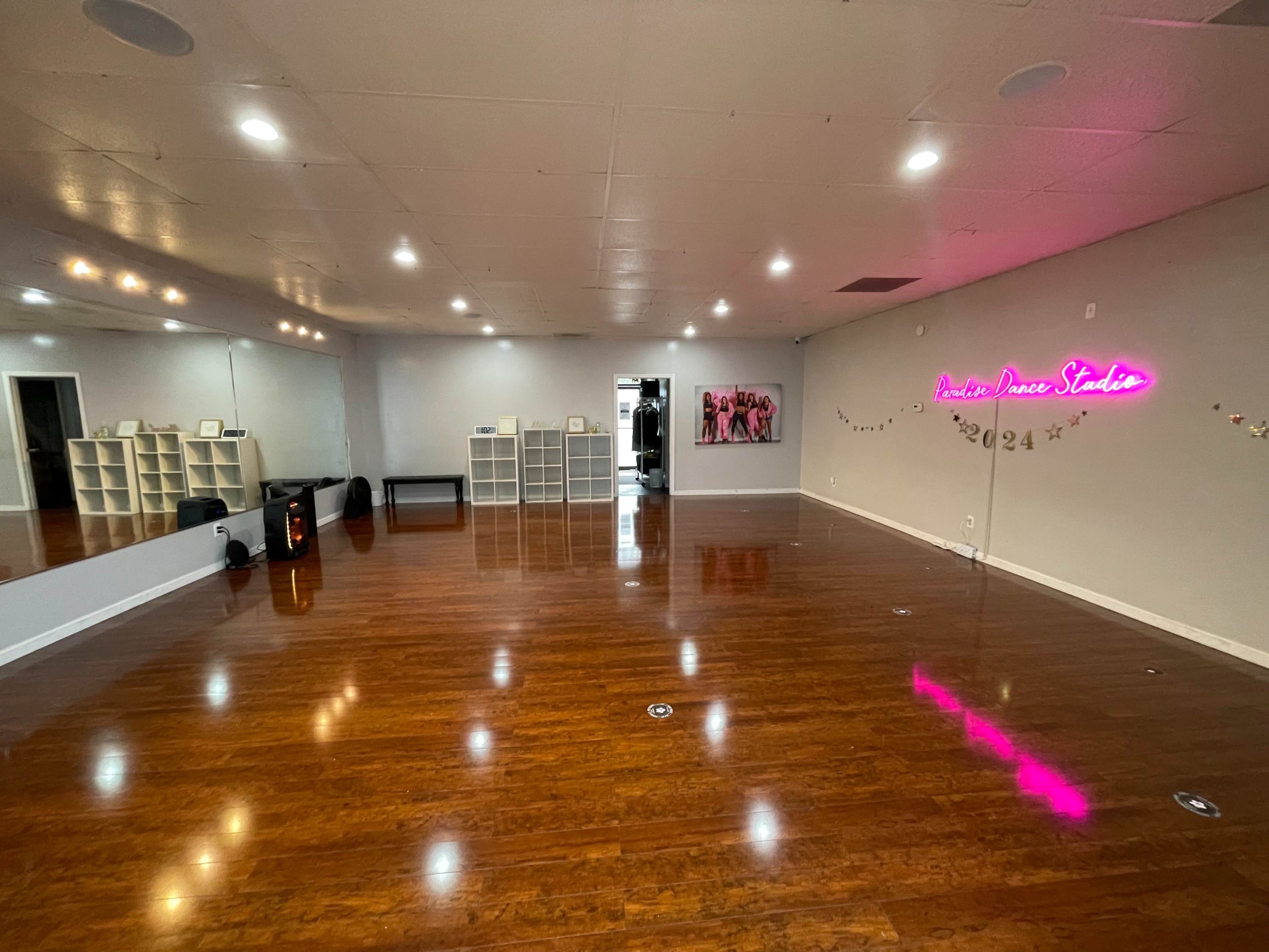 The image shows an empty dance studio with wooden flooring, wall mirrors, and a neon sign.