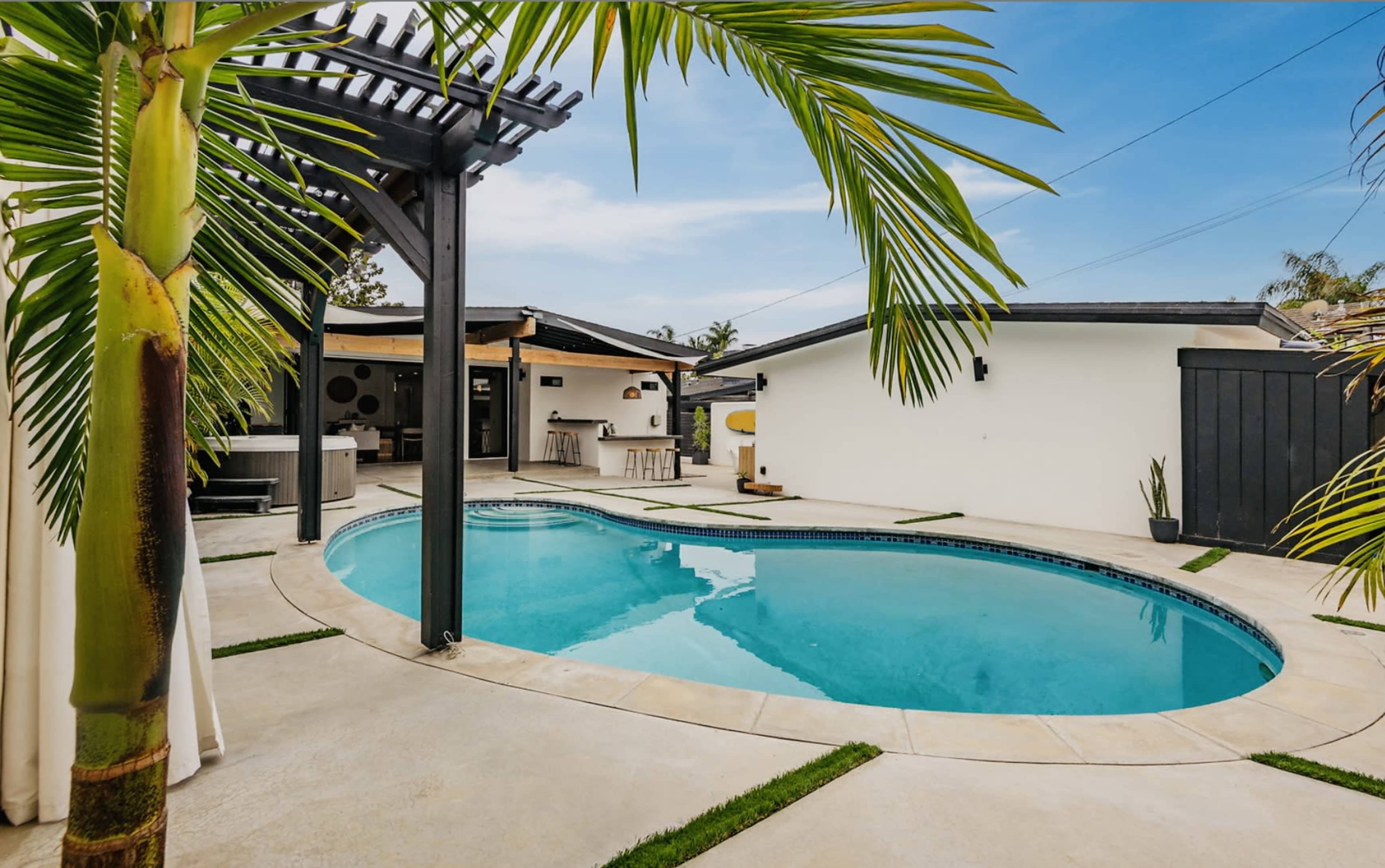 A curved swimming pool is surrounded by a concrete deck and palm trees, with a modern outdoor living area visible in the background.