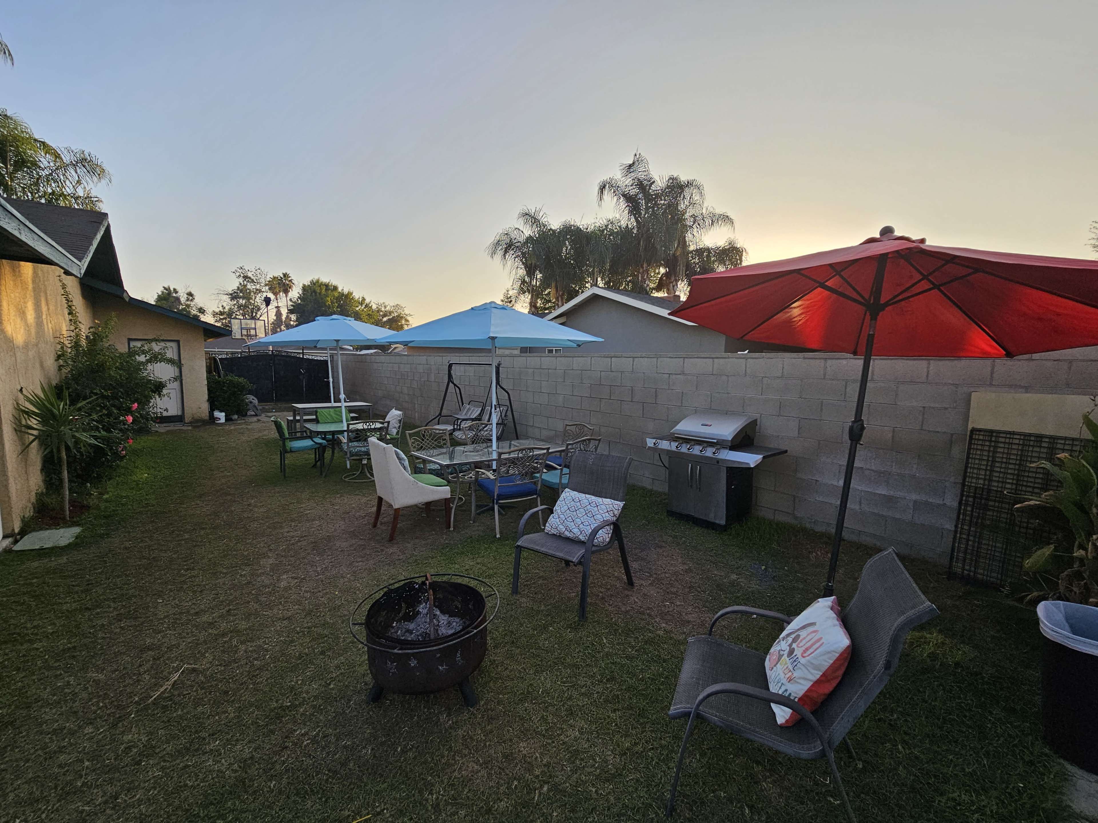 A backyard scene features several seating areas with umbrellas, a barbecue grill, and a fire pit surrounded by grass.