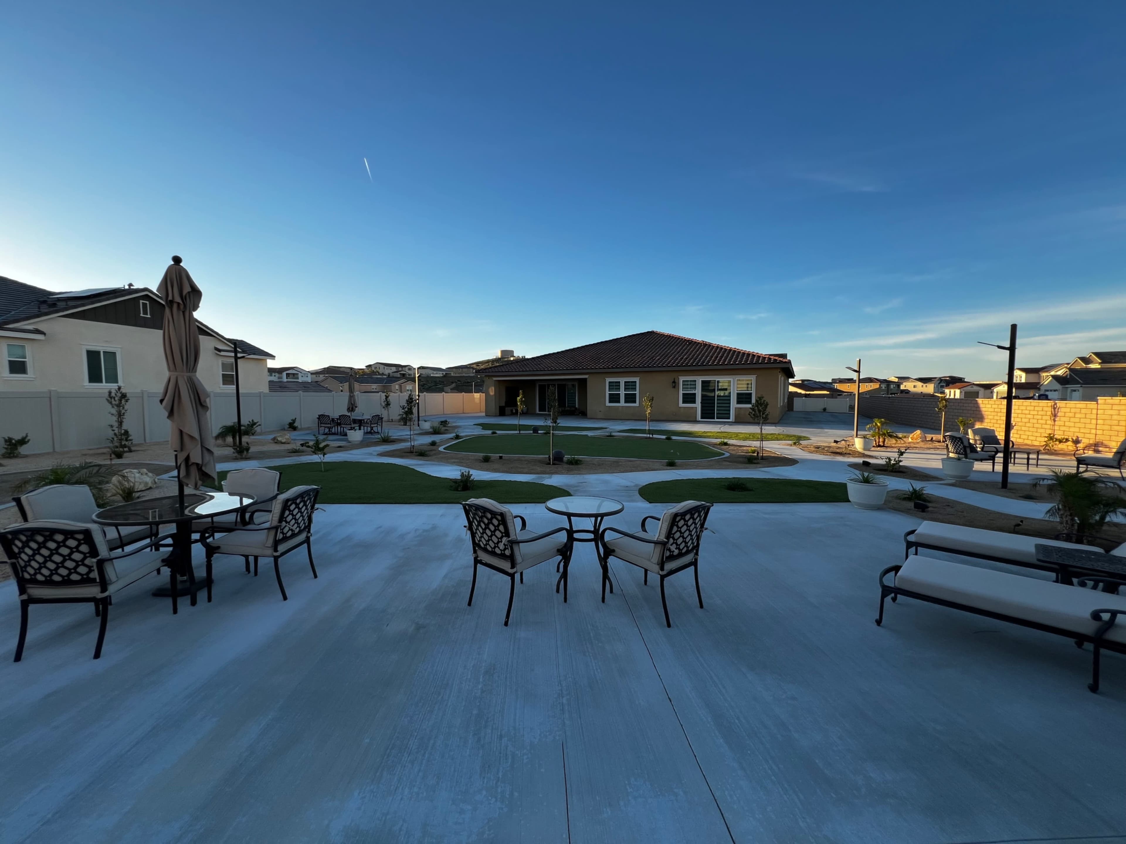 The image shows a landscaped backyard with seating areas, a lawn, and a yellow house in the background under a clear blue sky.