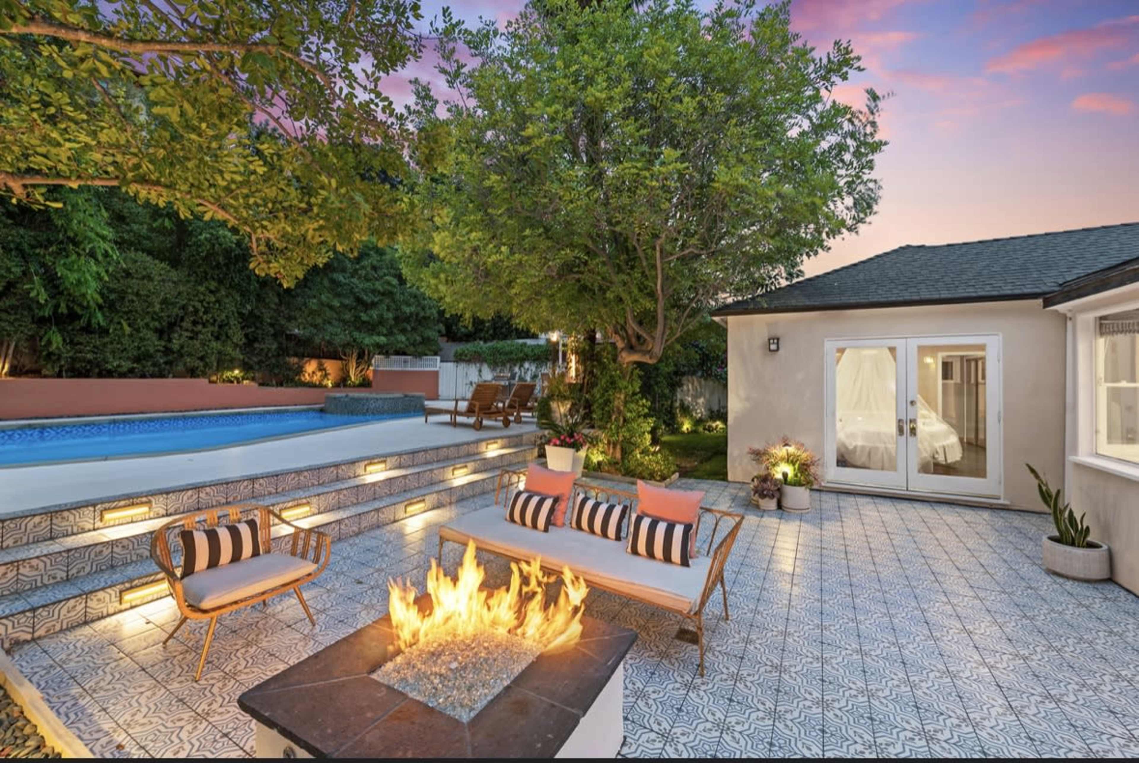 A backyard patio features a fire pit surrounded by seating, a swimming pool, and a cozy outdoor space with lush greenery.