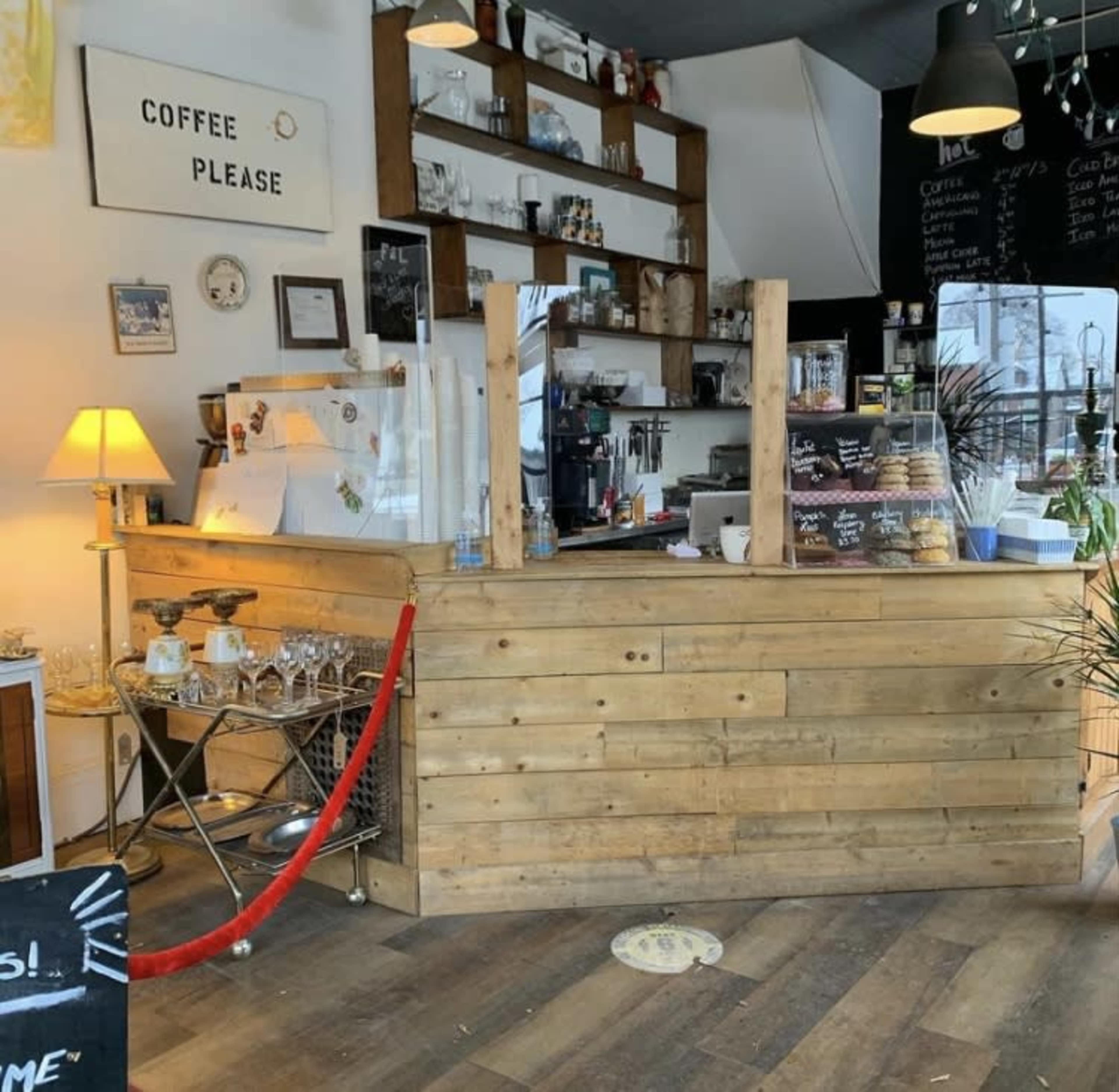 A wooden coffee shop counter with a glass display, shelves holding drinks, and a sign that says "Coffee Please."