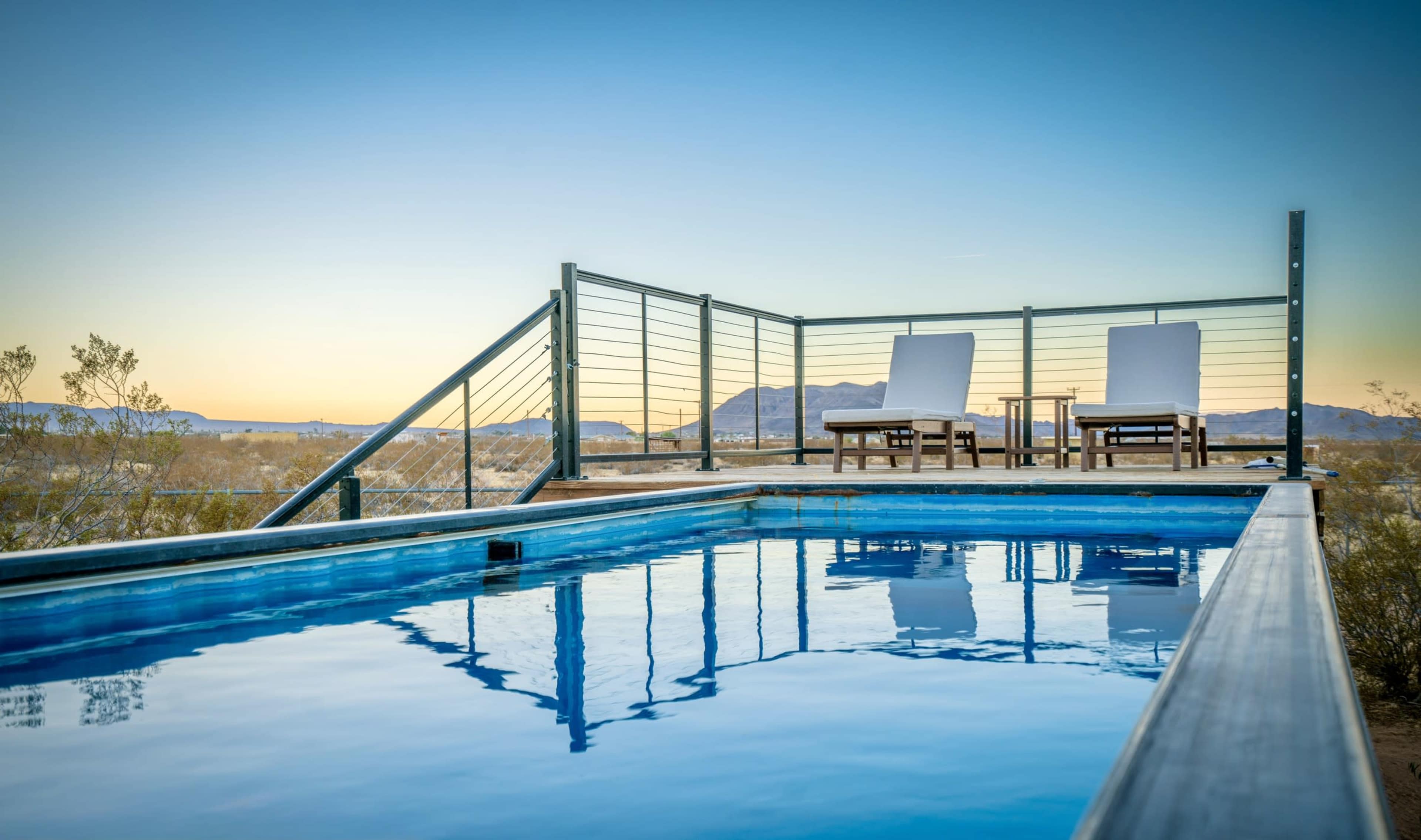 The image shows a rooftop swimming pool with two white lounge chairs beside it, surrounded by a desert landscape under a clear sky at dusk.