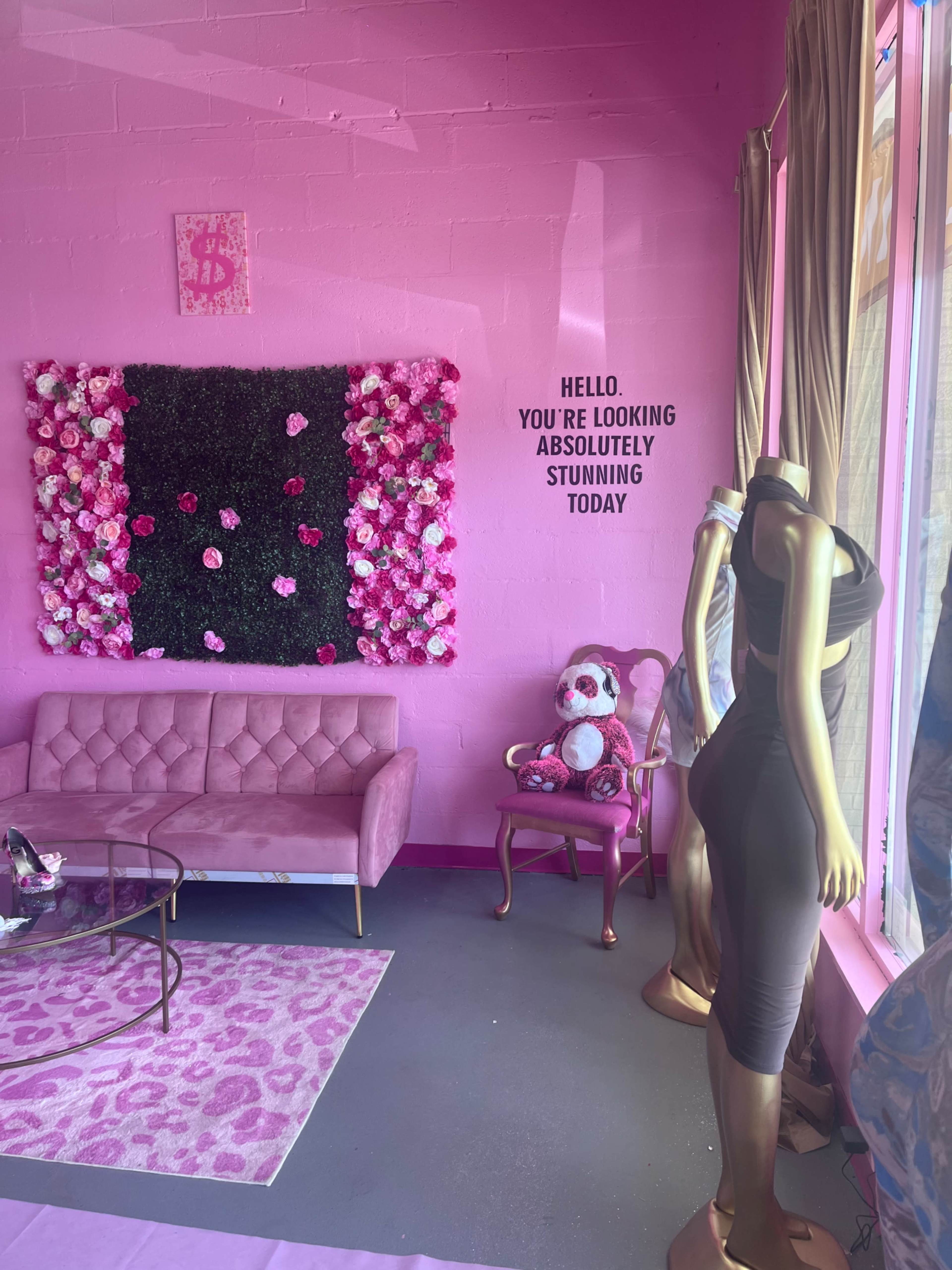 The image shows a brightly colored pink room with a floral wall decoration, a pink couch, a decorative teddy bear, and a mannequin displaying a dress.