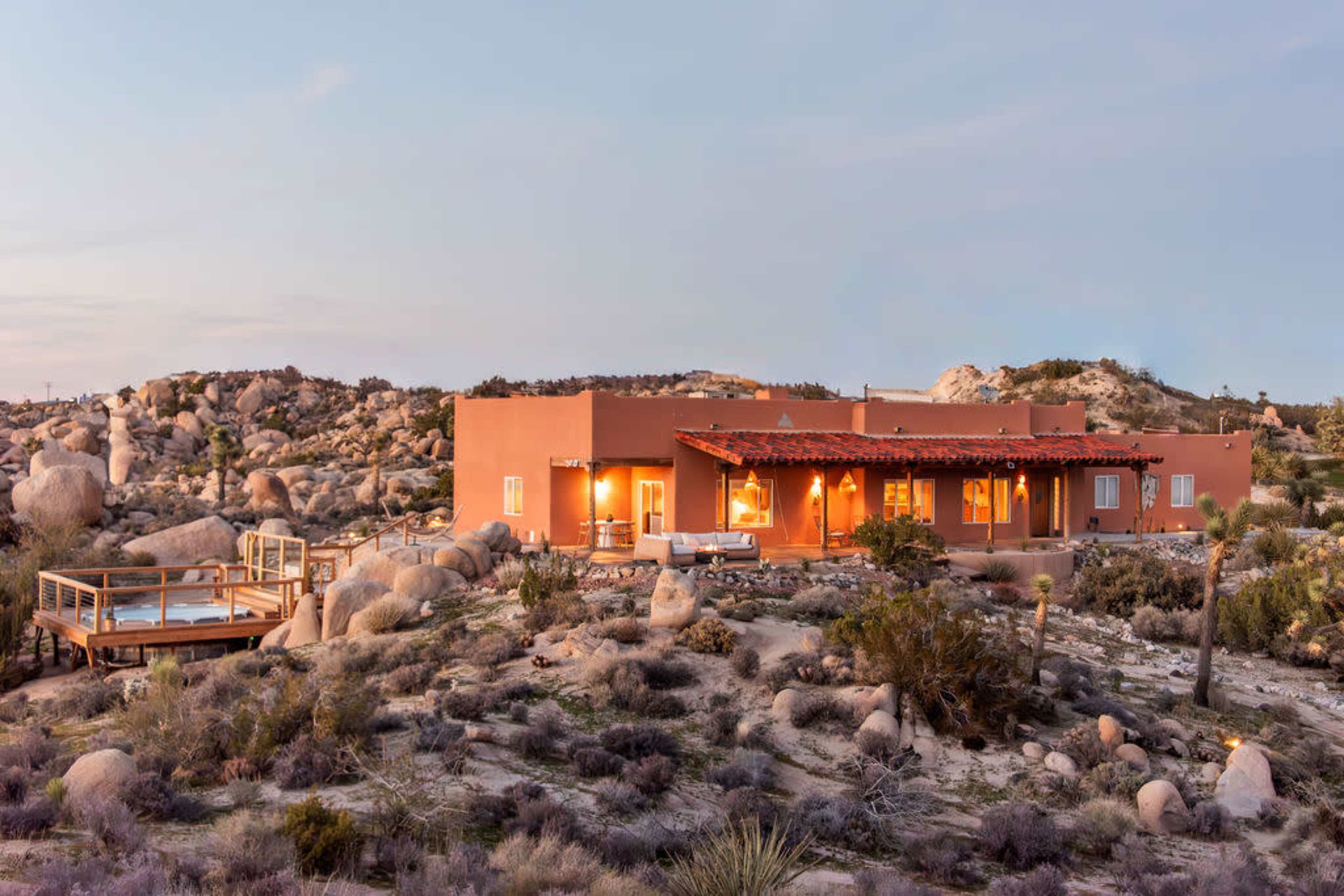 A single-story adobe-style house sits among large boulders and desert vegetation at dusk, illuminated by soft lighting from the windows.