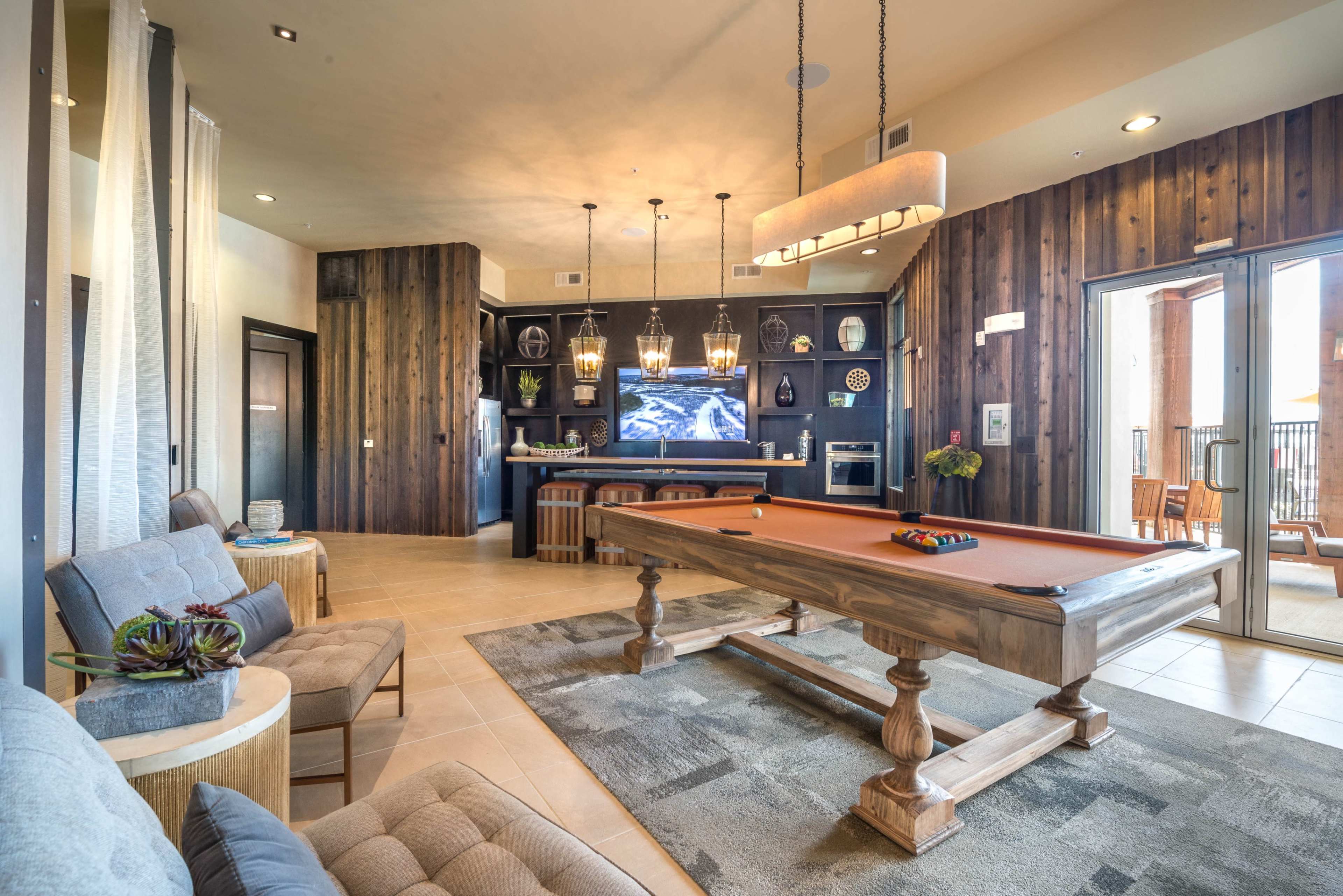 The image shows a spacious game room featuring a pool table, modern seating, and a kitchenette area with a large TV on the wall.