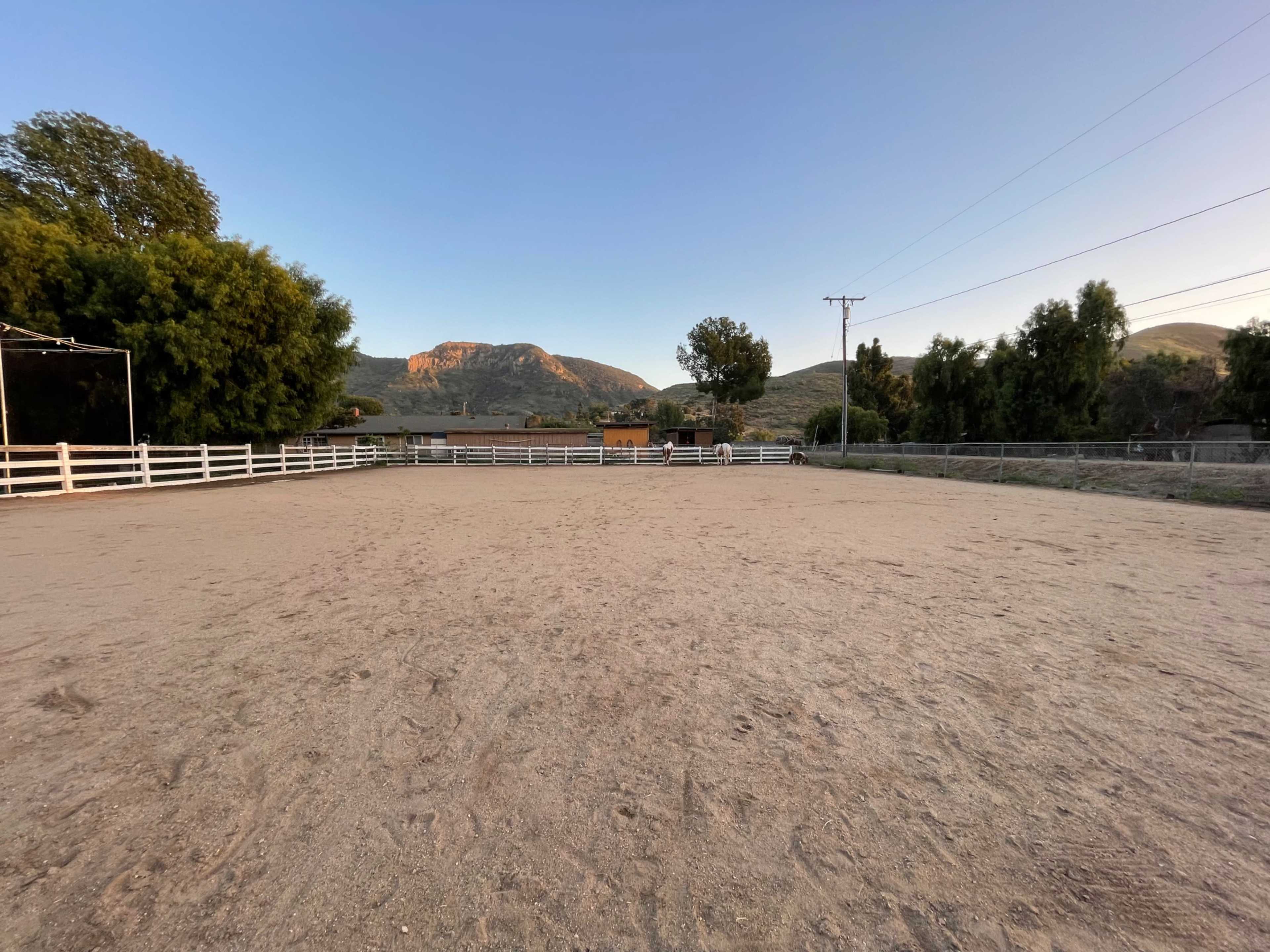 A sandy outdoor riding arena surrounded by trees and hills under a clear sky.