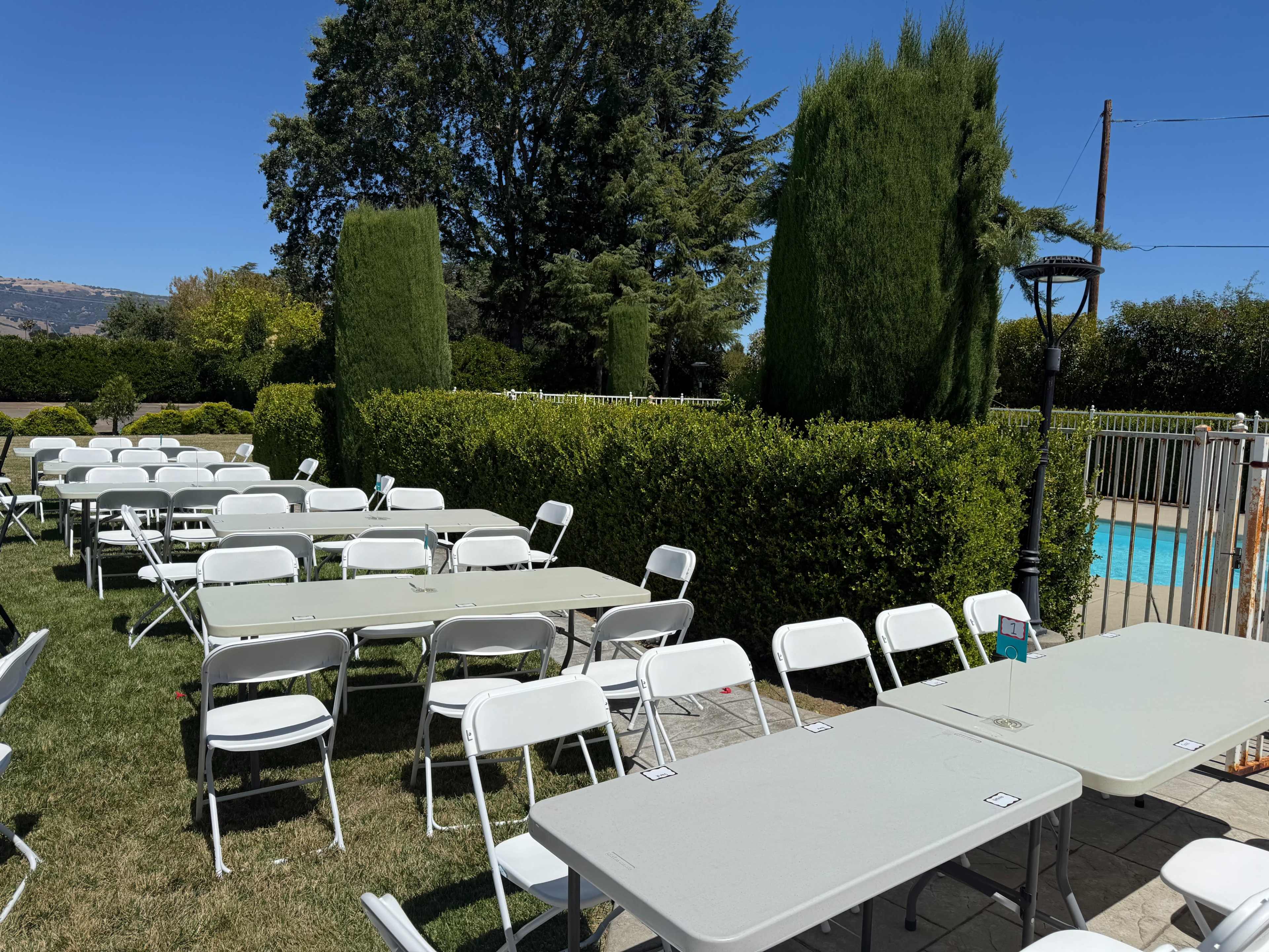 A row of white folding tables and chairs are set up on a lawn near a swimming pool, surrounded by neatly trimmed hedges and trees.