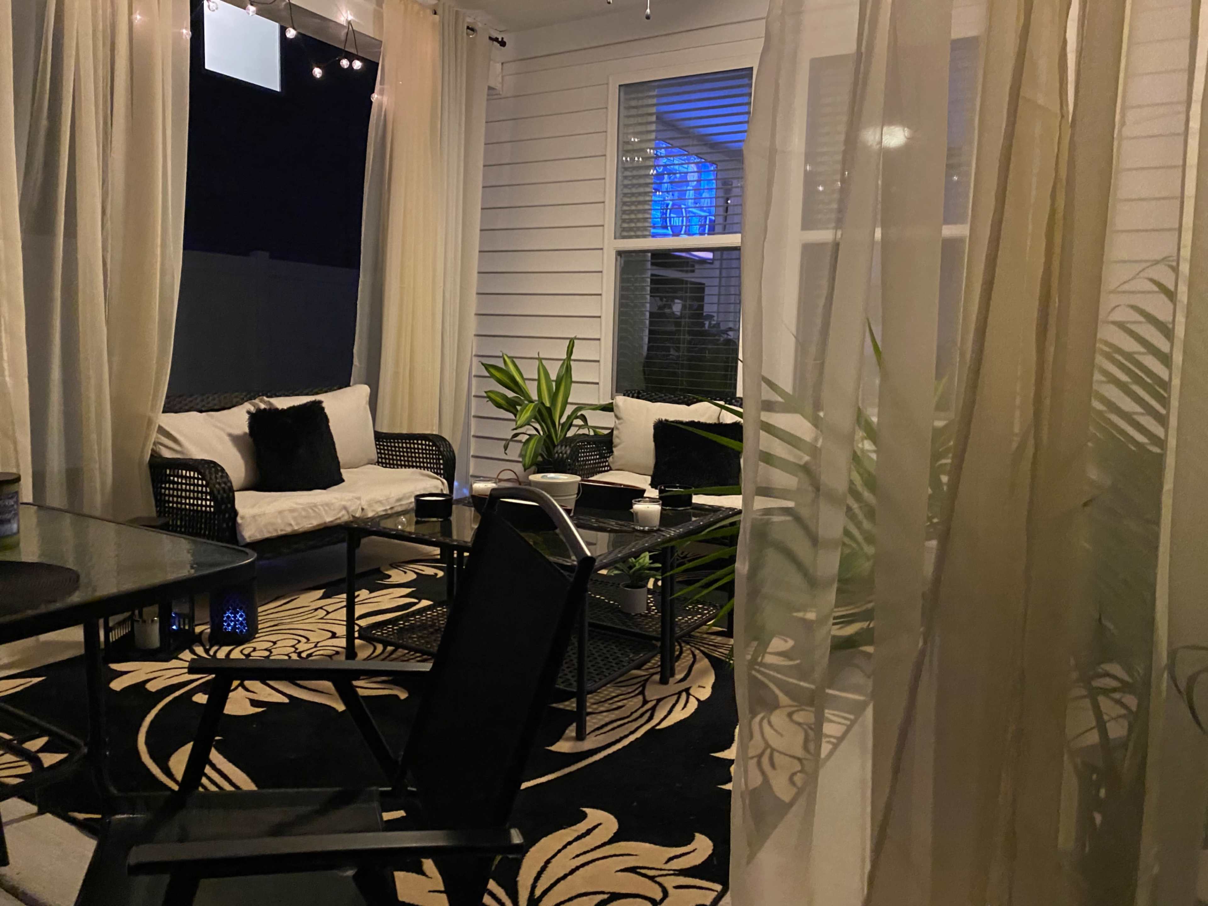 A patio area with black furniture, a patterned rug, and sheer curtains, illuminated softly at night.