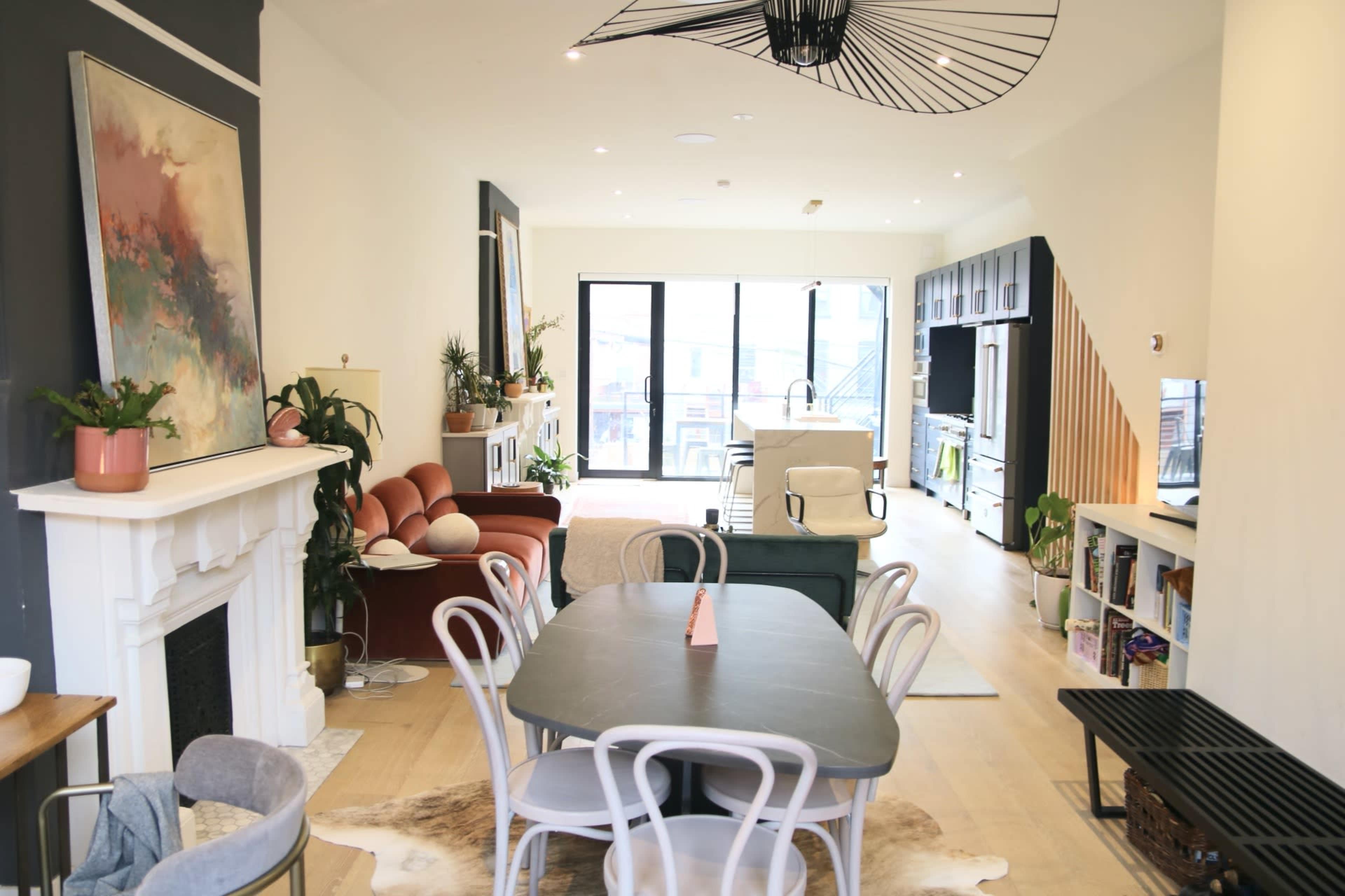 The image shows a contemporary open-plan living and dining area with a large window, featuring a mix of seating arrangements, plants, and a fireplace.