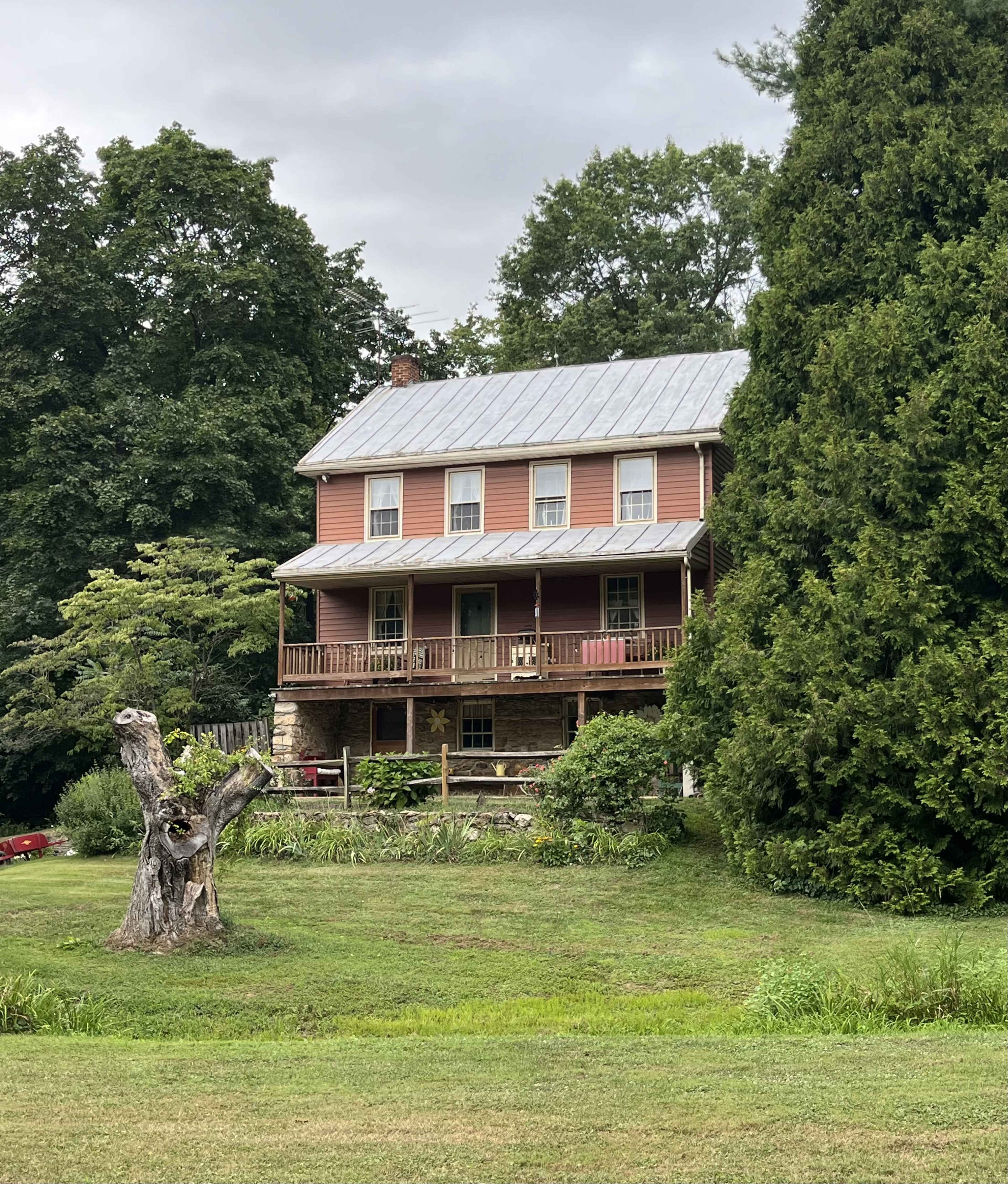 A two-story house with a metal roof and a wraparound porch sits amidst trees and greenery, alongside a weathered tree stump in the yard.