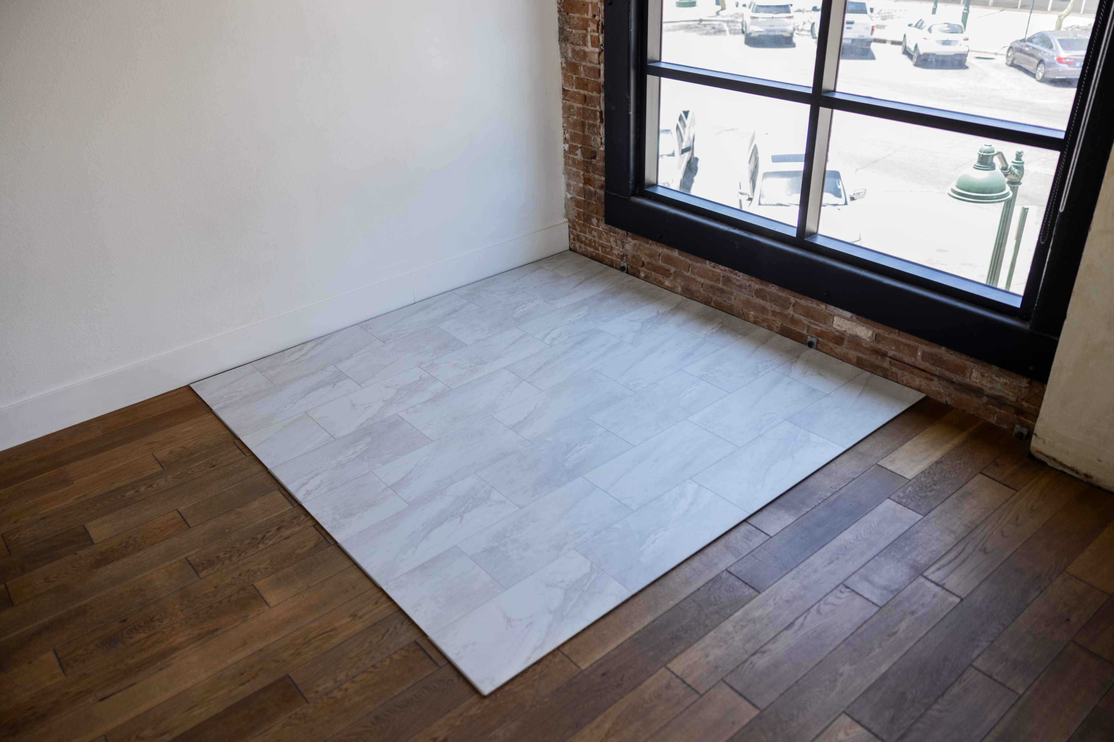 A corner of a room featuring a section of light-colored tile flooring next to darker hardwood flooring, with a large window providing daylight.