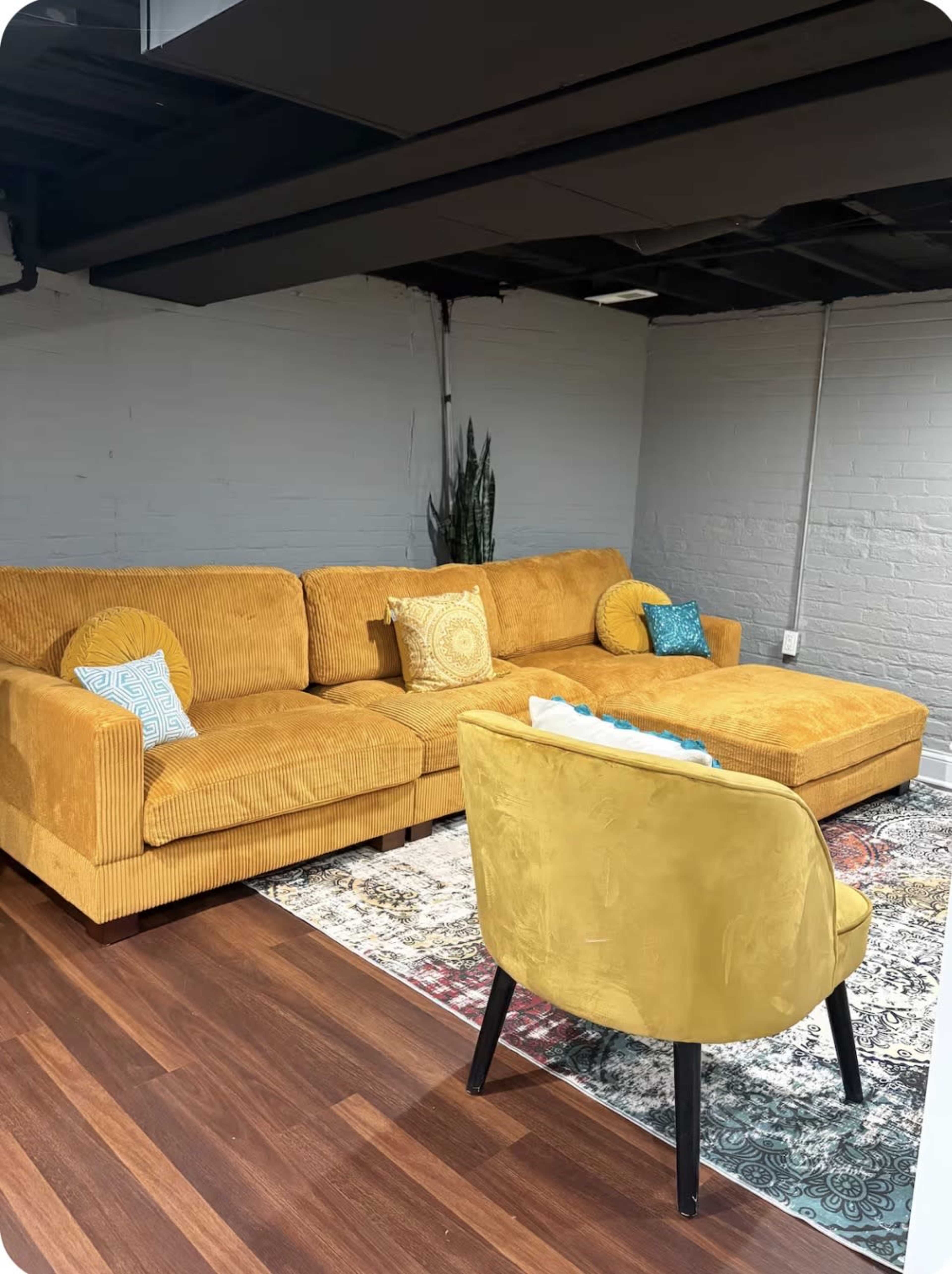 The image shows a living room featuring a large mustard yellow corduroy sectional sofa, a patterned area rug, and a single mustard yellow chair, set against a backdrop of gray-painted walls and a dark ceiling.