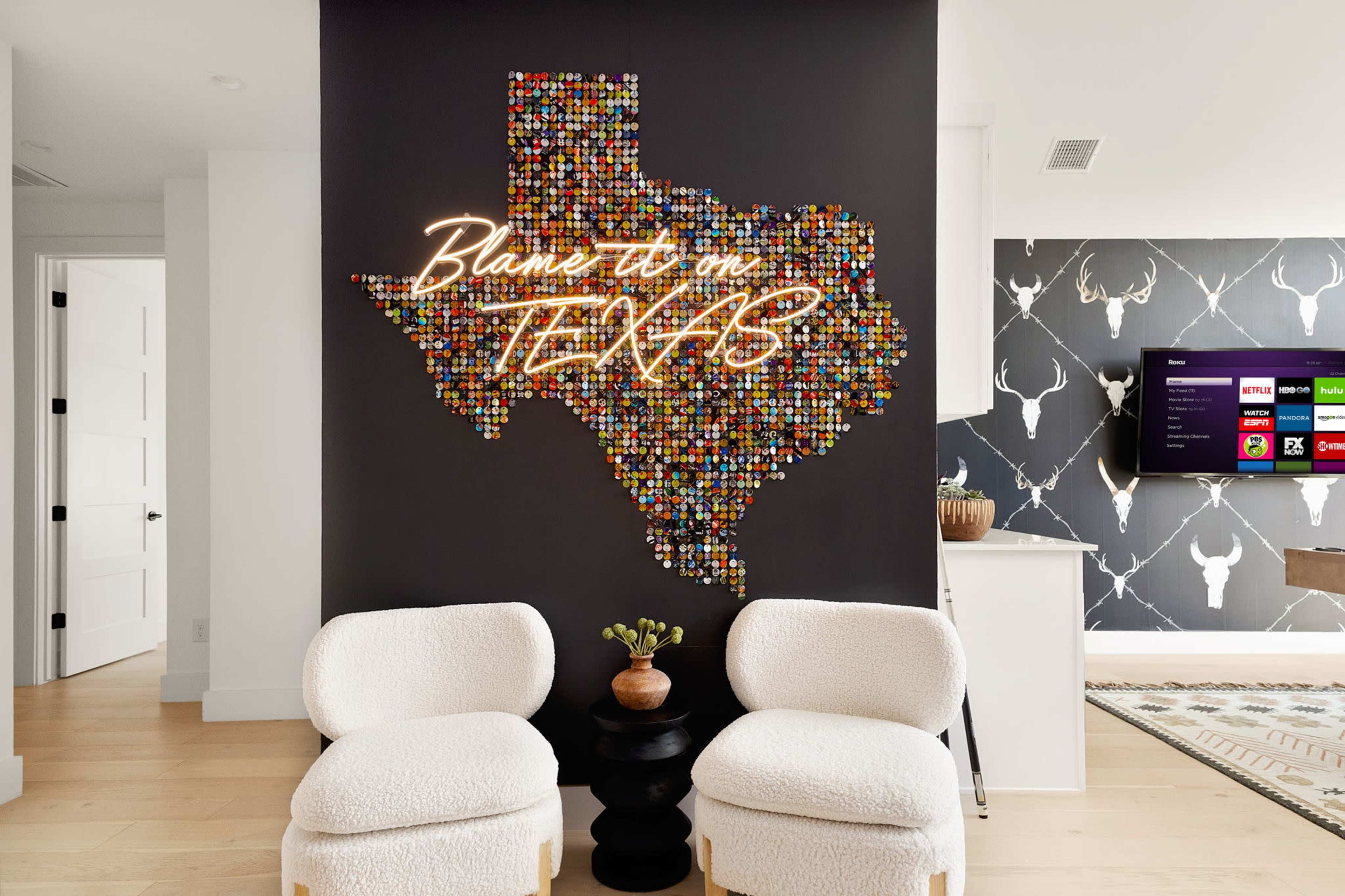A large Texas-shaped artwork made of colorful bottle caps is displayed on a black wall, accompanied by a neon sign that reads "Blame it on Texas," with two white armchairs in the foreground.