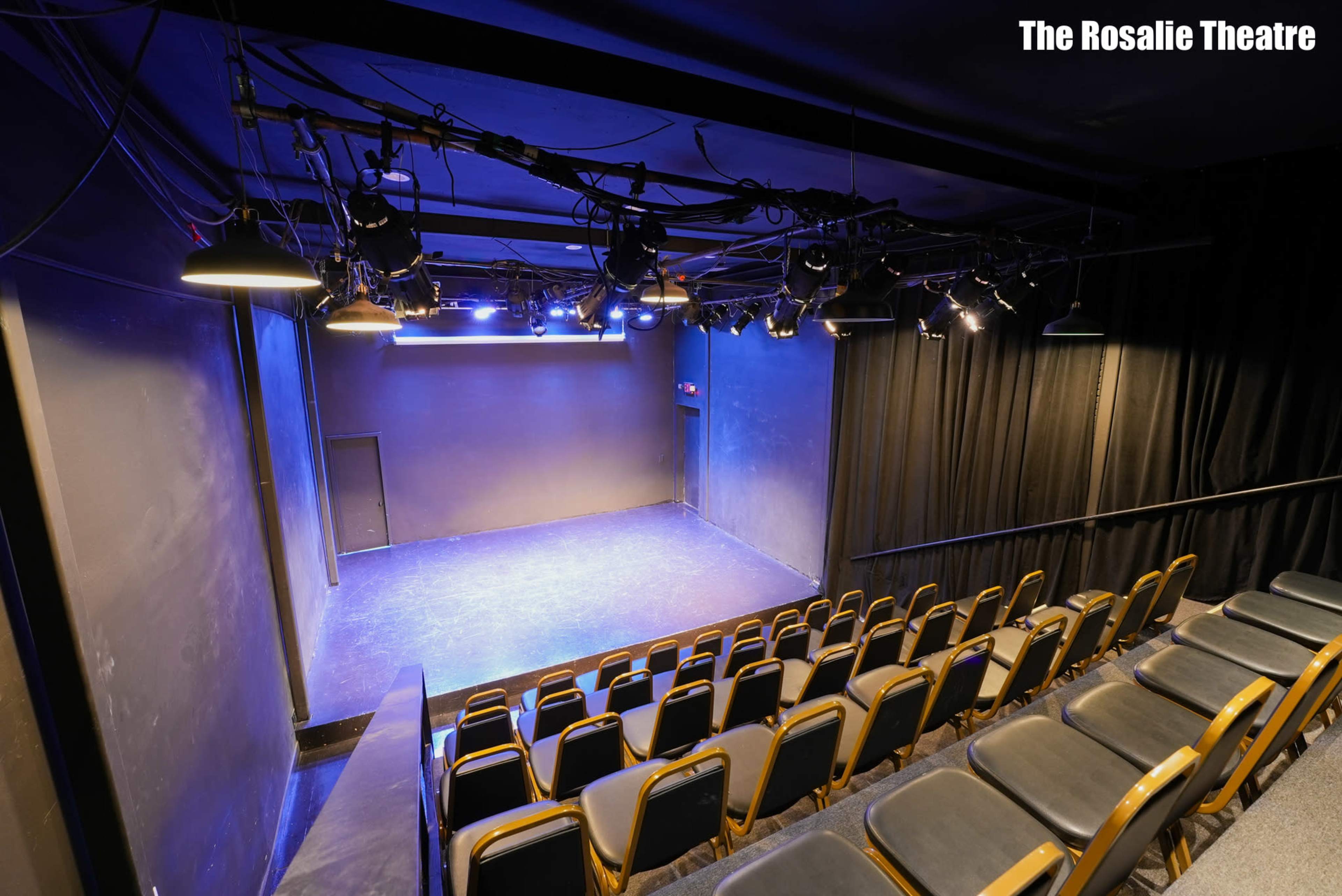 The image shows an empty theater with black walls, rows of seatings facing a small stage illuminated by overhead lights.