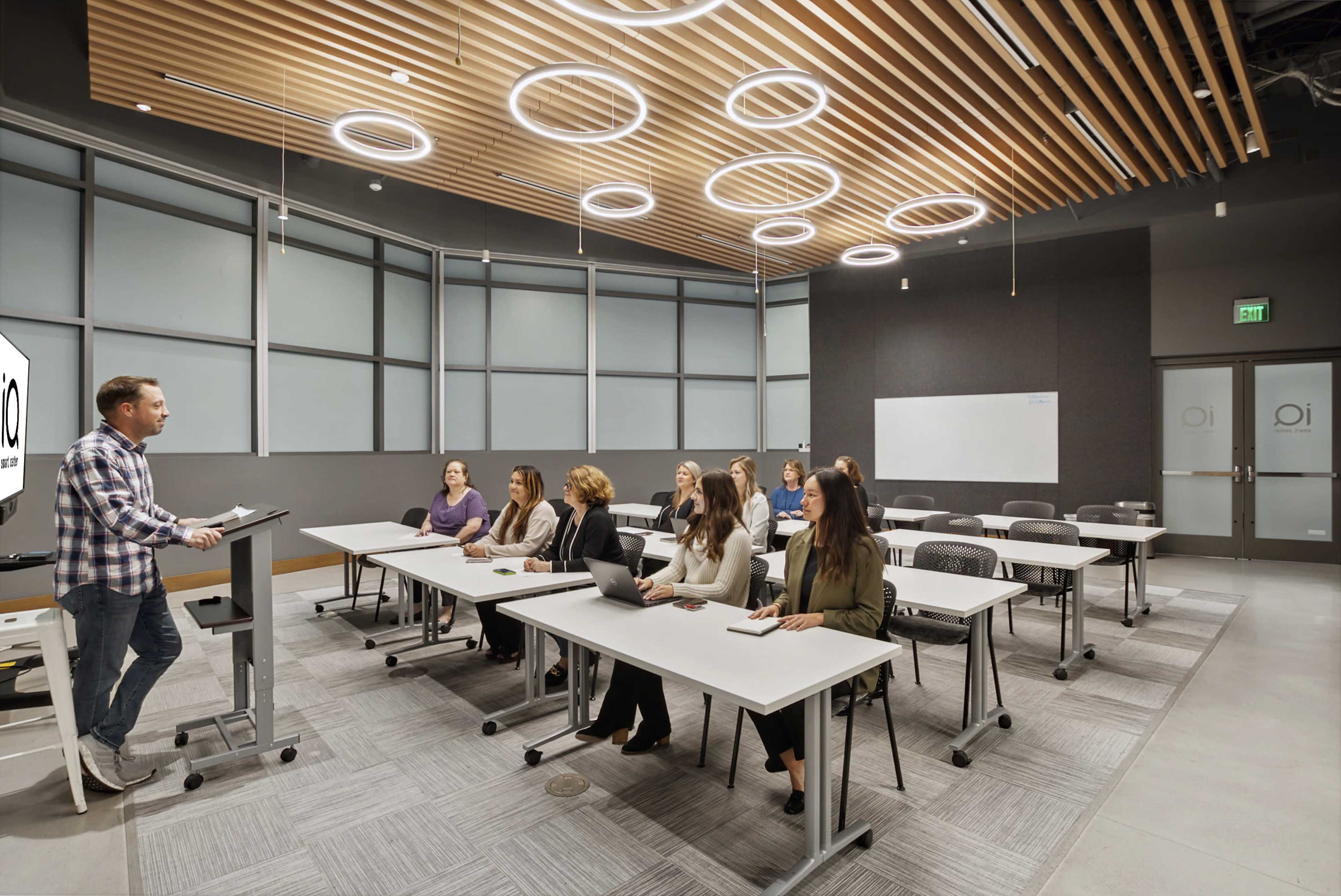 A man gives a presentation to a group of seated participants in a modern classroom with circular ceiling lights and a flexibly arranged seating layout.