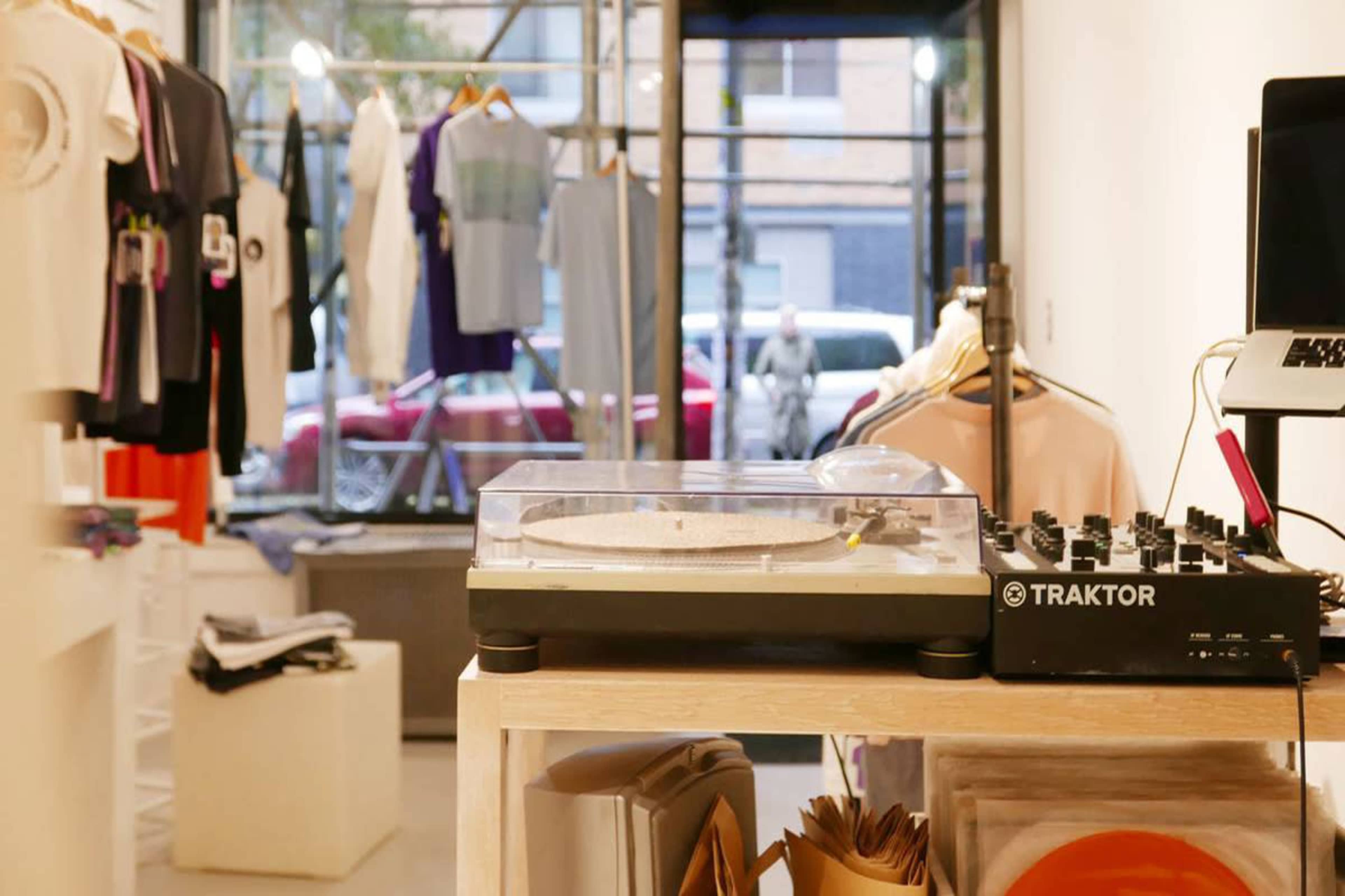 A DJ setup with a turntable and a mixer on a wooden table, surrounded by clothing displayed on racks in a store.