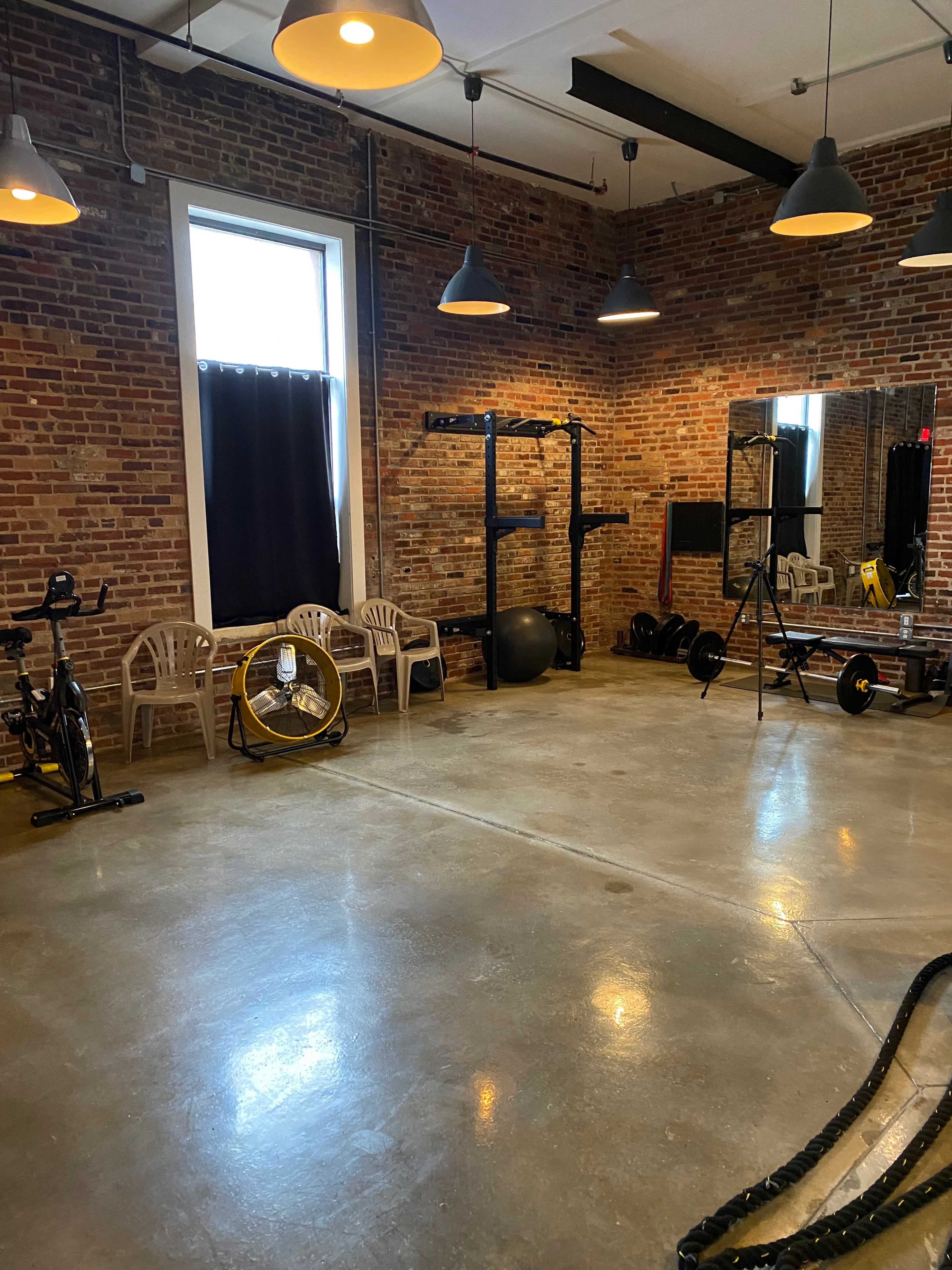 A minimally furnished gym with exposed brick walls, a large mirror, exercise equipment, and several plastic chairs.