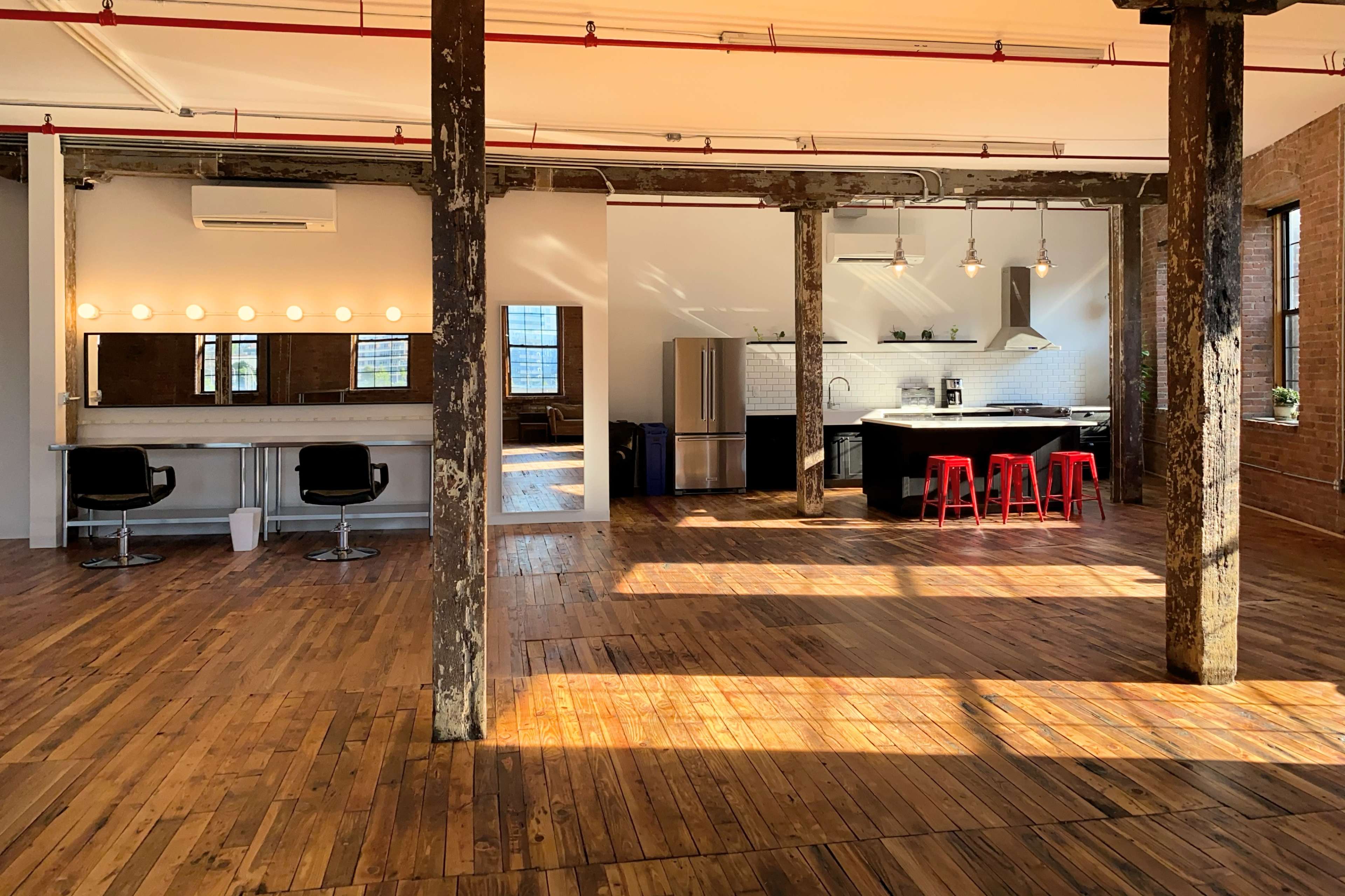The image shows a spacious, industrial-style interior with wooden floors, exposed pillars, and a kitchen area featuring stainless steel appliances and red bar stools.
