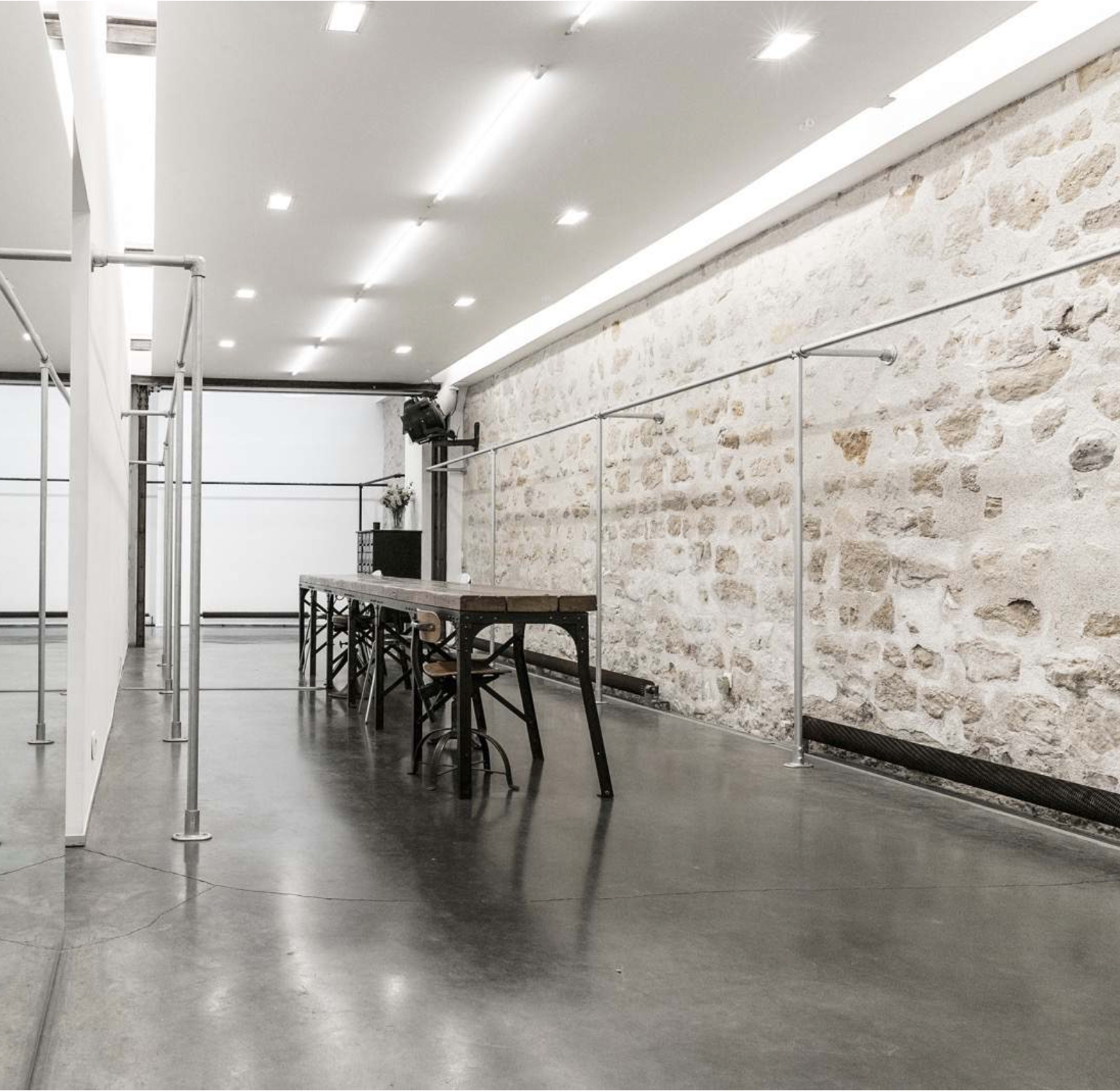 A spacious interior features a long wooden table surrounded by chairs, with a stone wall and minimalist lighting.