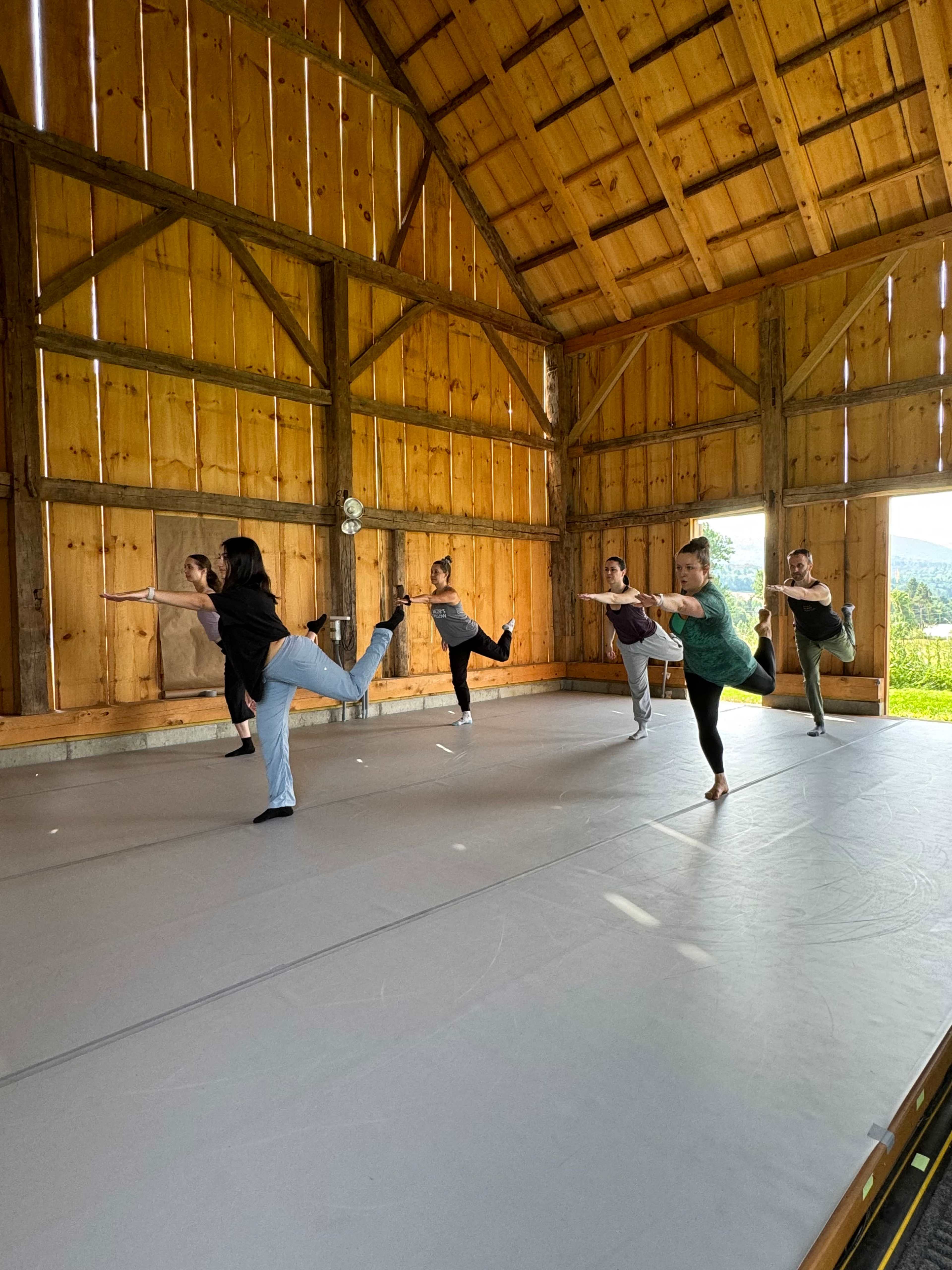 A group of six dancers practices in a spacious, wooden barn-style studio with a large open wall and natural light coming through.