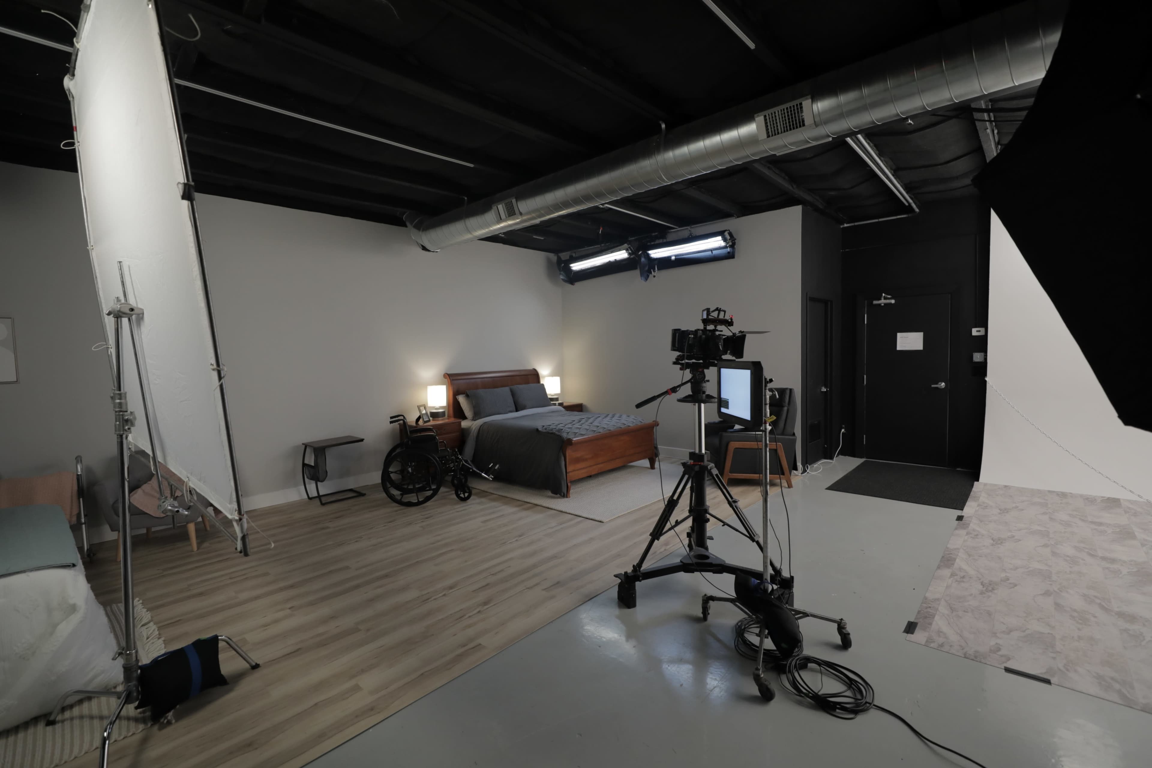 A bedroom setup for a photo or video shoot, featuring a bed, a wheelchair, and several lighting and camera equipment arranged on a light-colored floor.