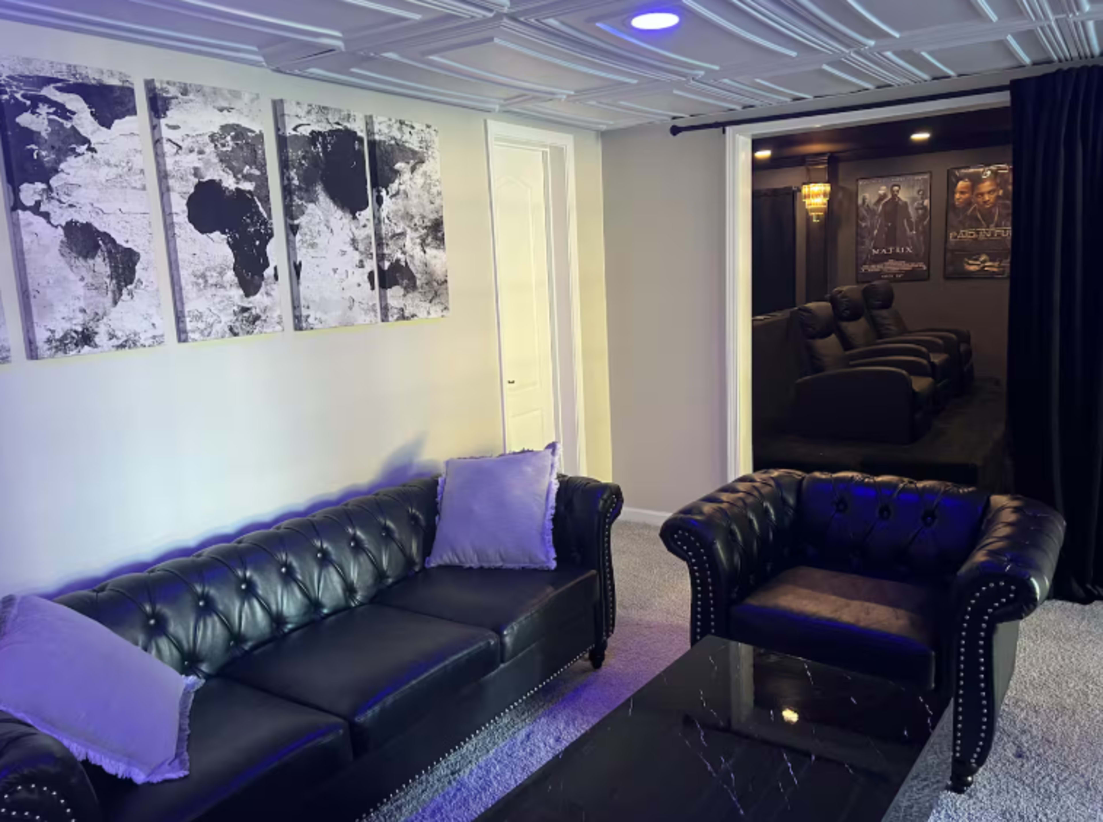 The image shows a modern lounge area with black leather sofas, a glass coffee table, and wall art depicting world maps, leading to a dimly lit movie room in the background.