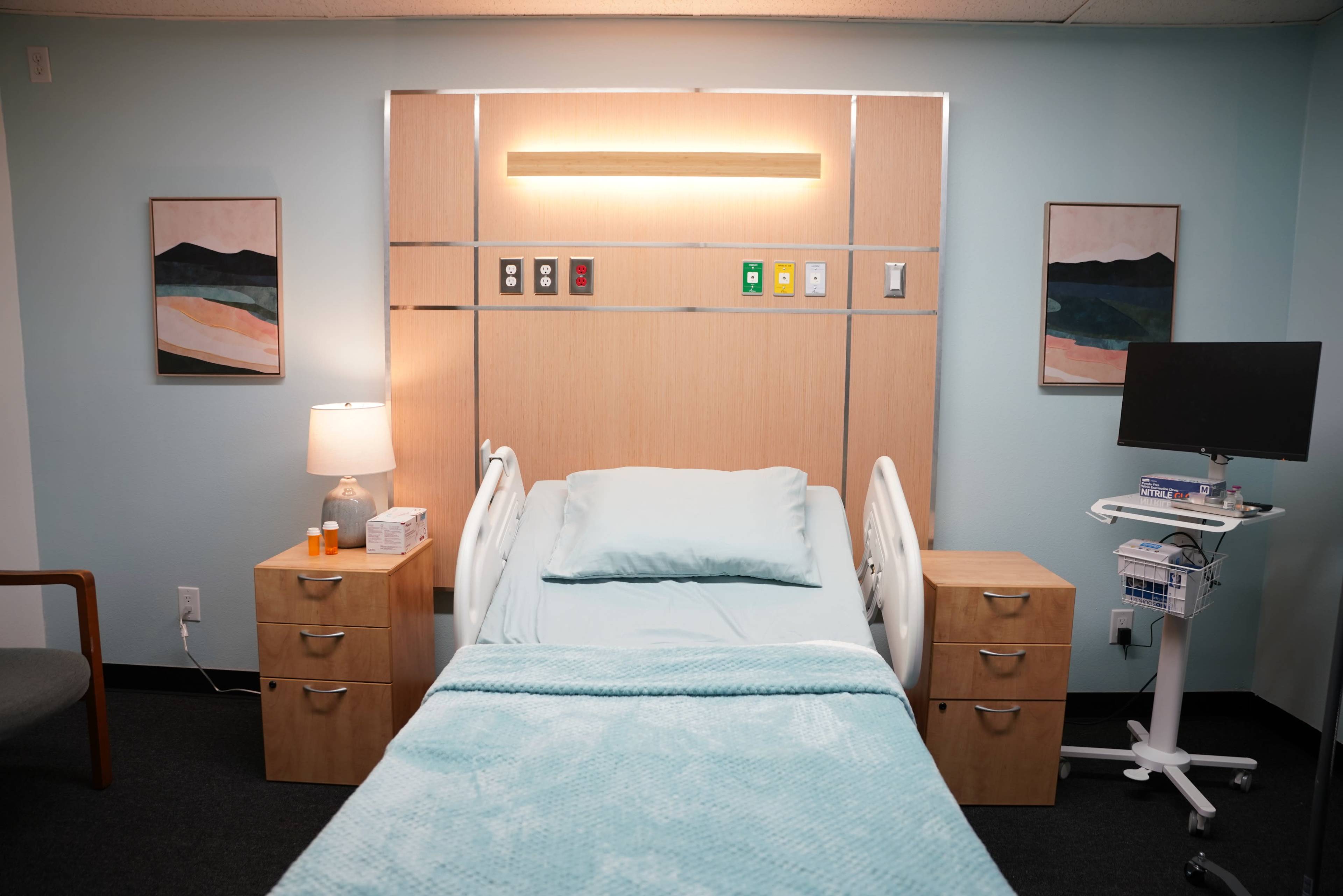 The image shows a hospital room featuring a patient bed with light blue bedding, flanked by wooden nightstands, a lamp, and a small medical cart with equipment.