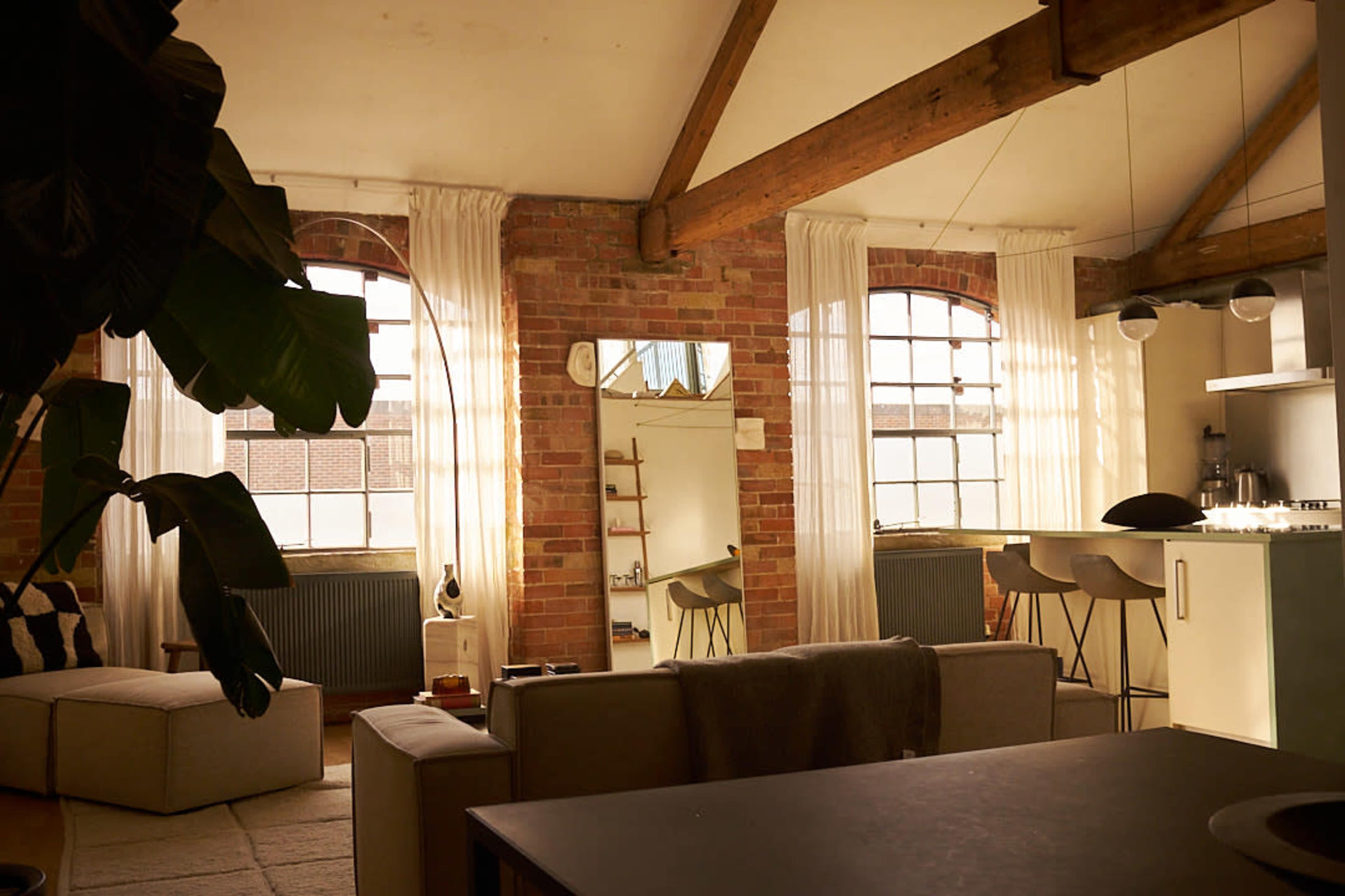 The image shows a spacious, open-concept living area featuring exposed brick walls, large windows, and modern furnishings.