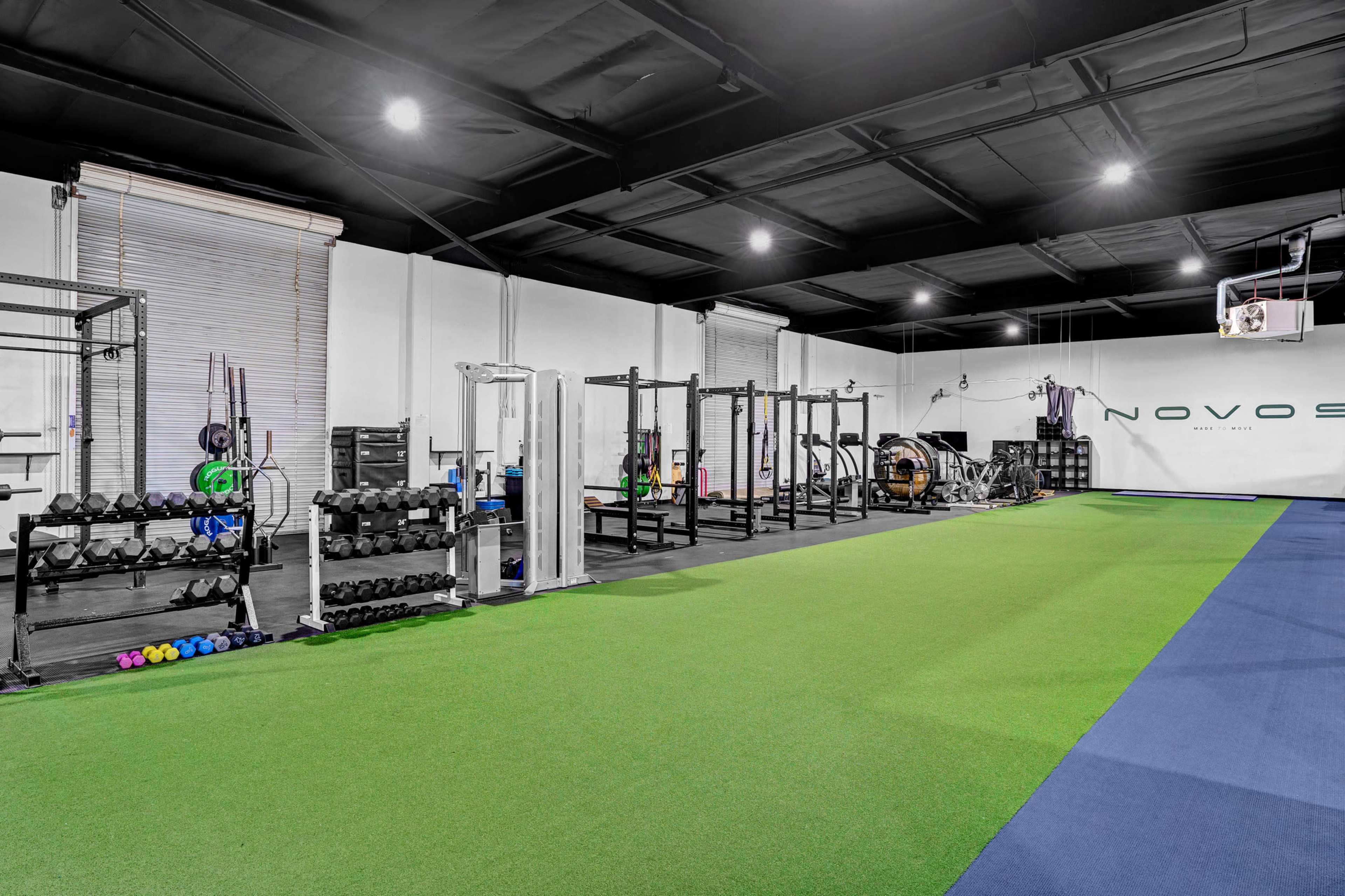 A gym interior with a green turf area, weightlifting equipment, and a wall featuring the name "NOVOS."