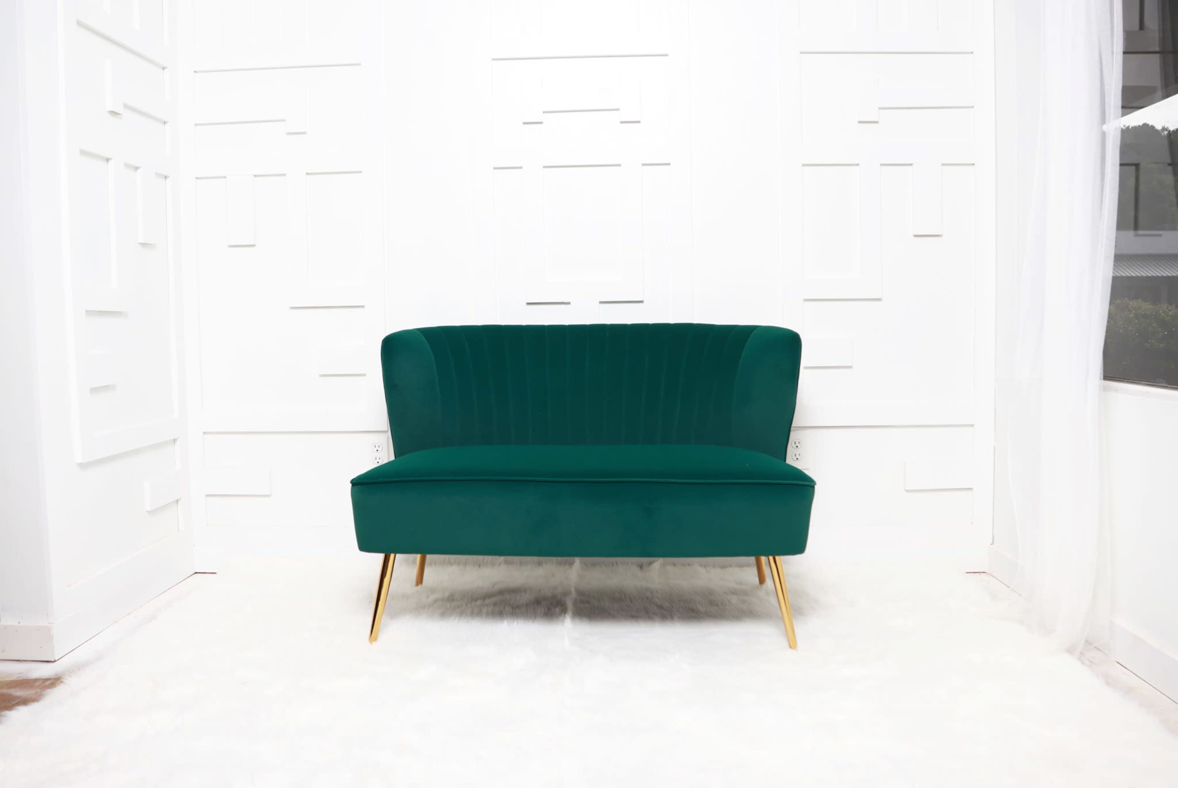 A green velvet loveseat with gold legs is positioned in a bright room with a white backdrop.