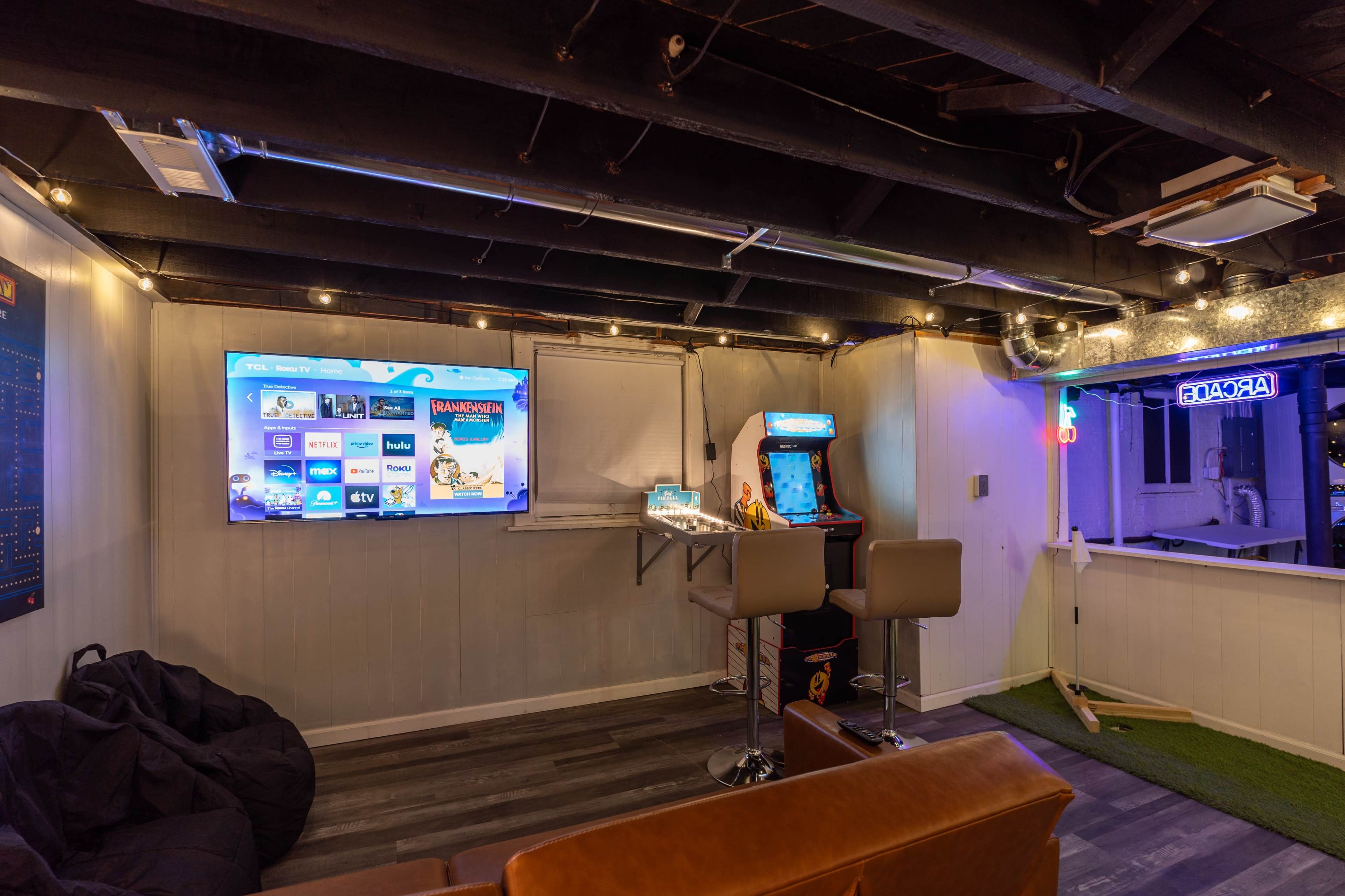 The image shows a modern game room featuring a large screen, an arcade cabinet, bar stools, and ambient lighting.