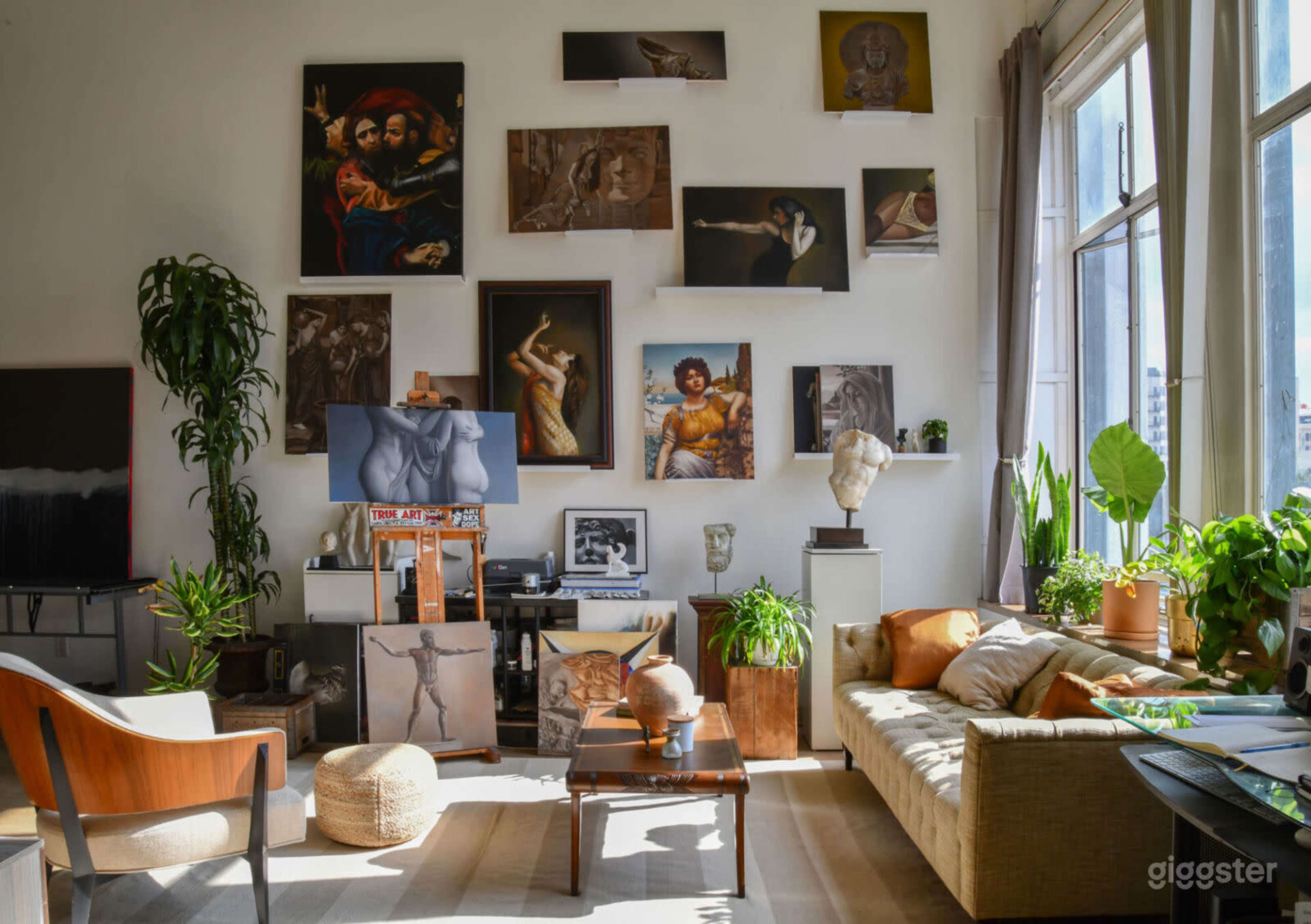 The image depicts a bright, artistic living space with various paintings on the walls, plants in the corners, and comfortable furniture arranged around a central coffee table.