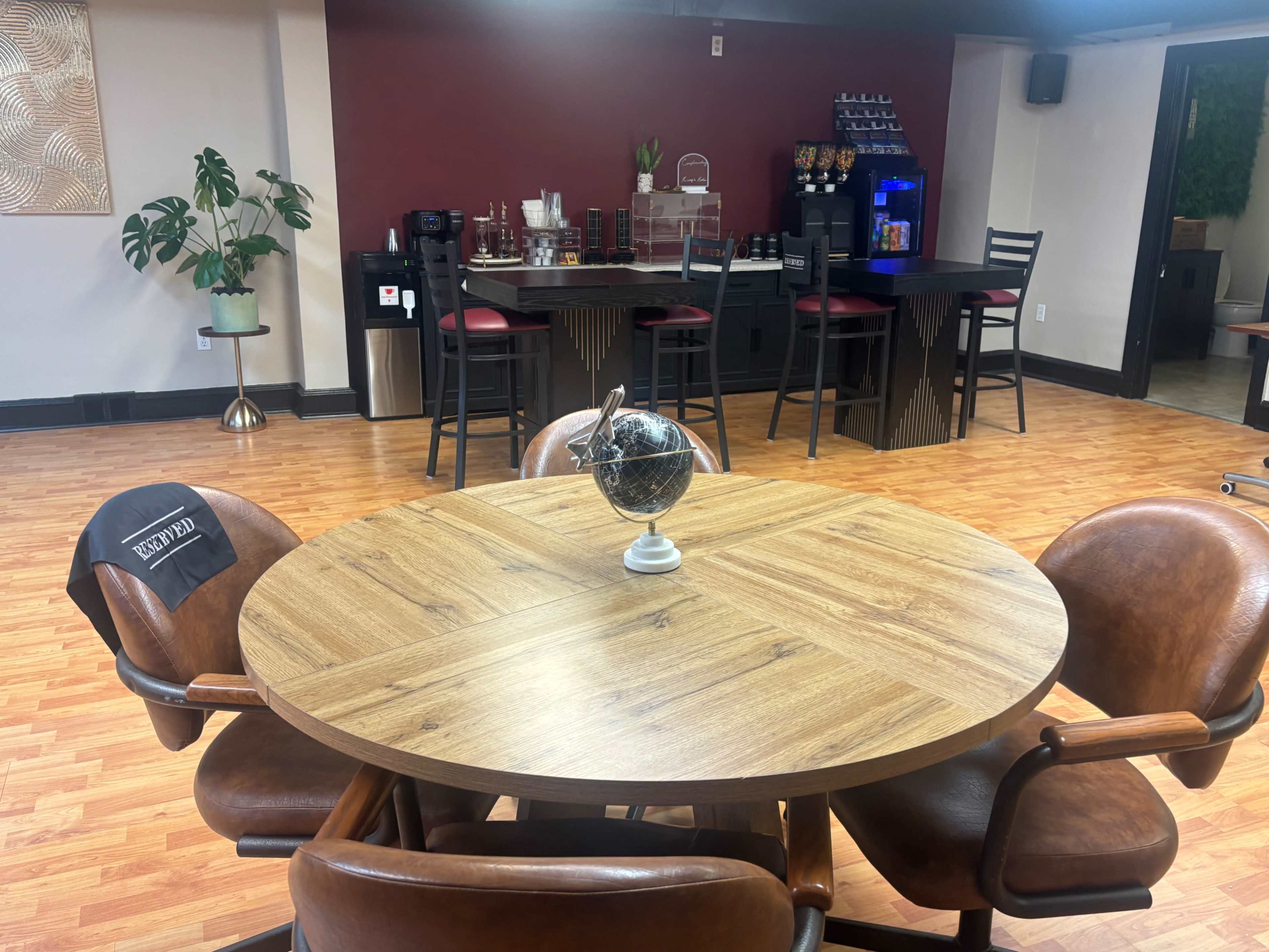 The image shows a spacious room featuring a round wooden table surrounded by leather chairs, with a bar counter and several high chairs in the background.